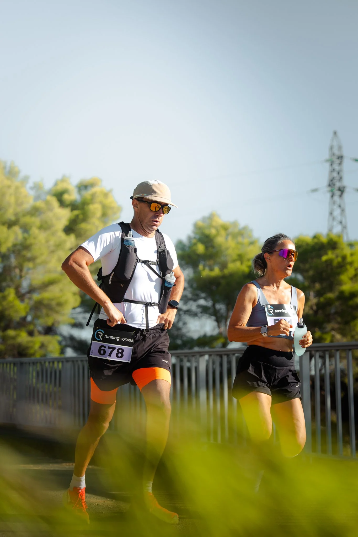 Deux coureurs en tenue de sport participant à une course, portant des numéros 678 et 57, dans un environnement extérieur avec des arbres et un pylône électrique en arrière-plan.