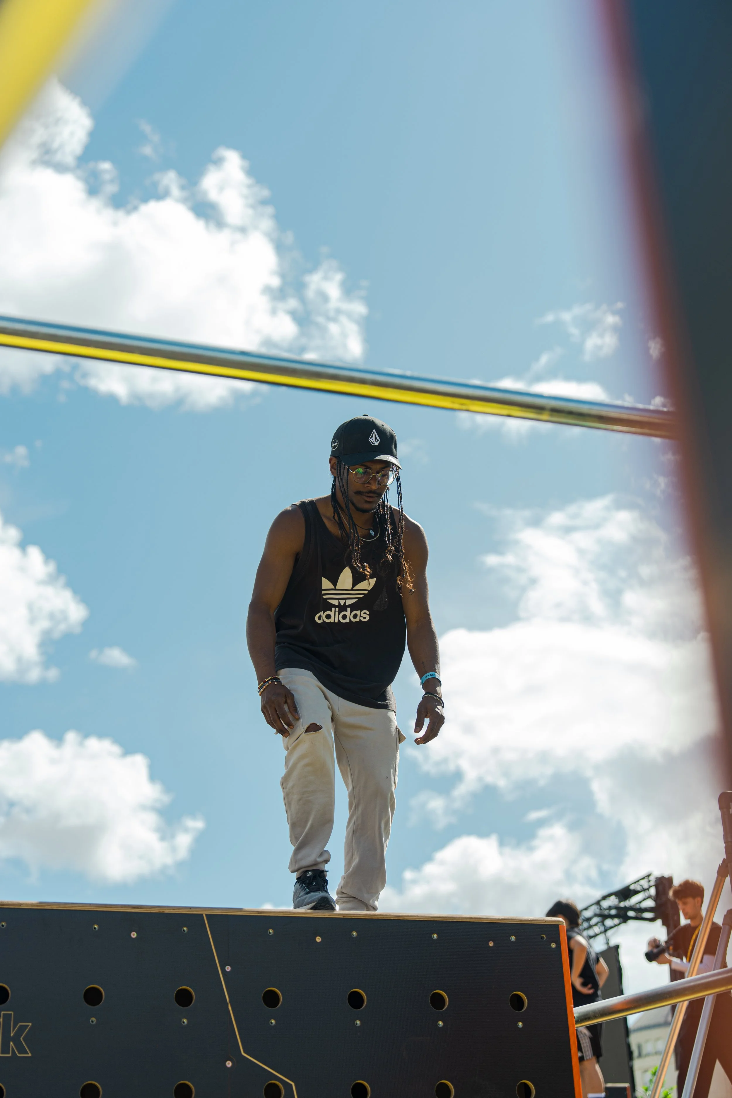 Une personne portant un t-shirt noir Adidas, des lunettes et une casquette, marche sur une plateforme lors d'un événement en plein air avec un ciel bleu et des nuages, plusieurs autres personnes en arrière-plan.
