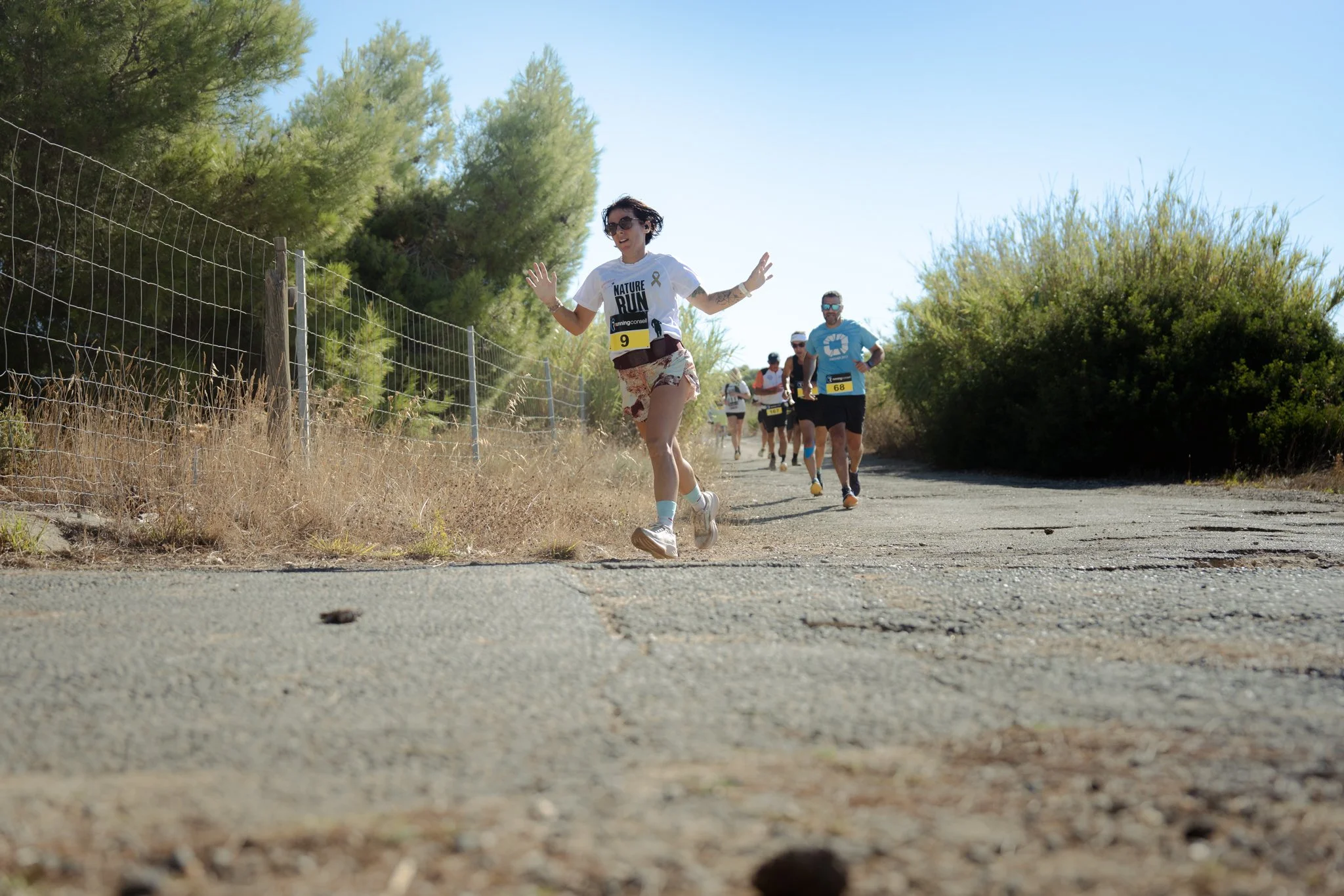 Des personnes participent à une course ou un marathon dans un environnement extérieur ensoleillé, avec des arbres verts et un ciel bleu. La femme en avant porte un t-shirt blanc avec l'inscription 'NATURE RUN'.