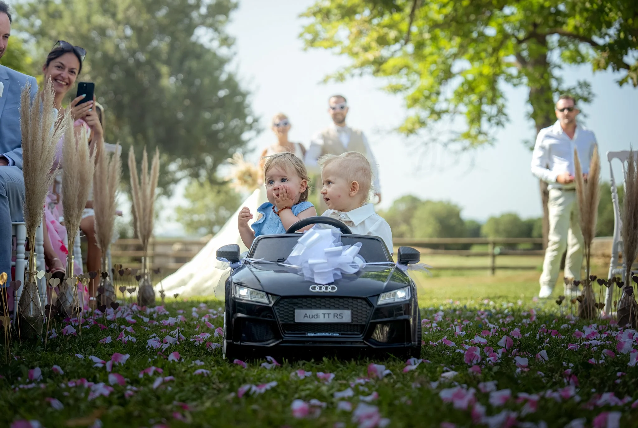 Une scène de mariage en plein air avec des invités souriants, un enfant dans une voiture miniature Audi TT RS décorée d'un nœud de mariage, entouré de fleurs rose et d'arbres en arrière-plan.