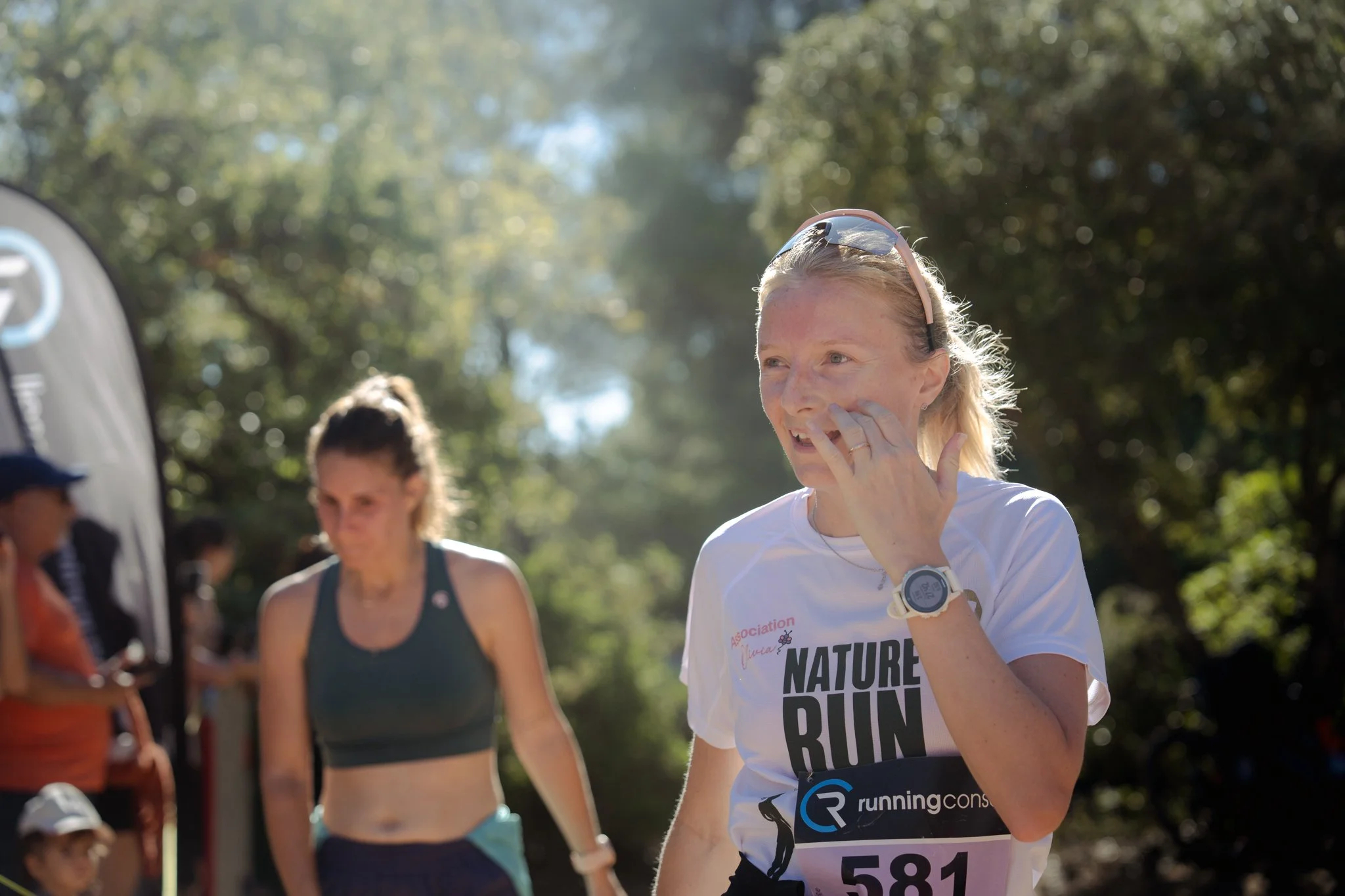 Femme couverte de sueur et souriante, portant des lunettes de soleil sur la tête, participant à une course dans un parc naturel avec d'autres coureurs en arrière-plan.