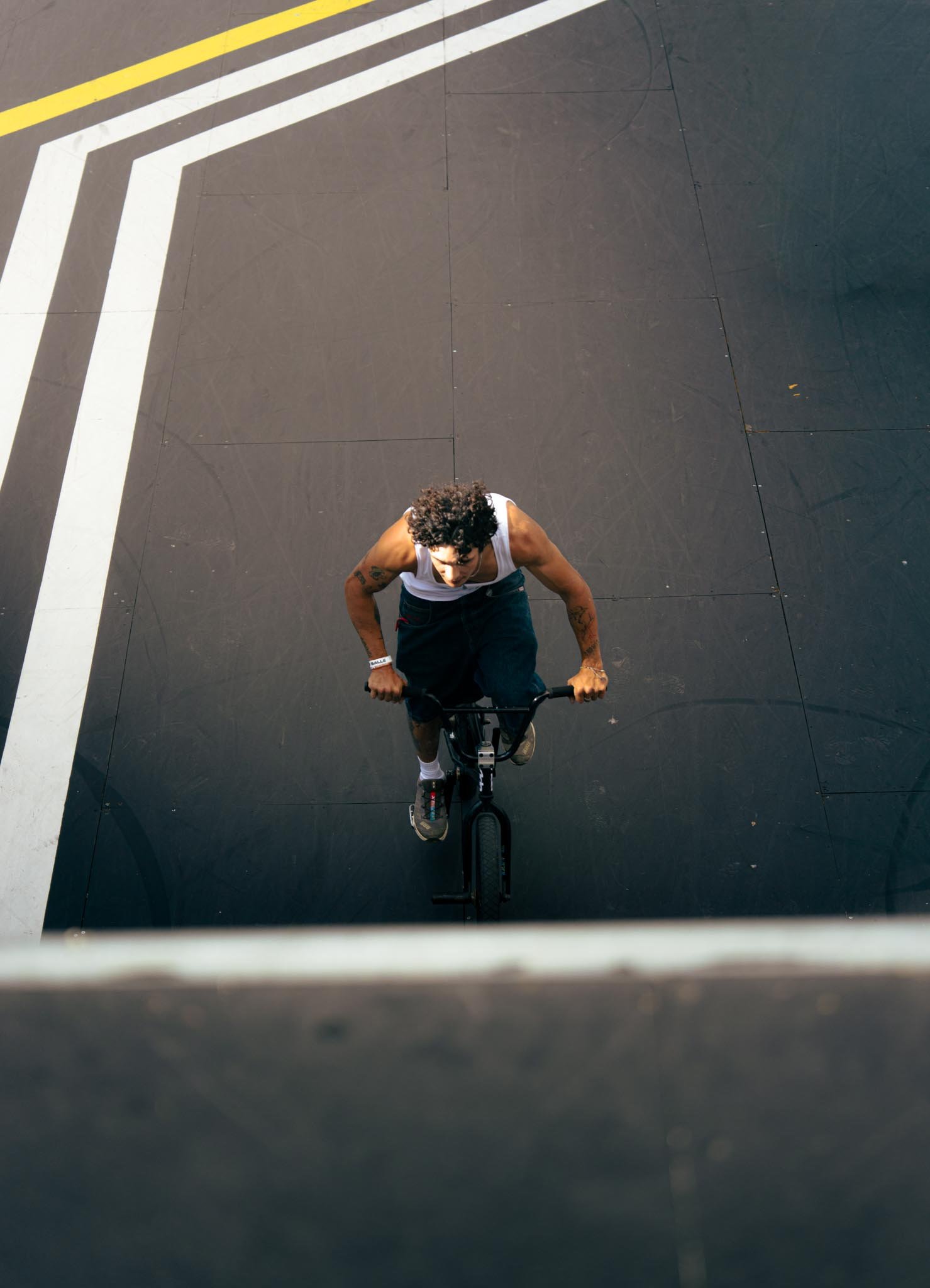 A person riding a black bicycle on a dark outdoor surface. The photo is taken from above, showing the person's curly hair, white tank top, and tattoos on their arms. White and yellow lines are visible on the pavement.