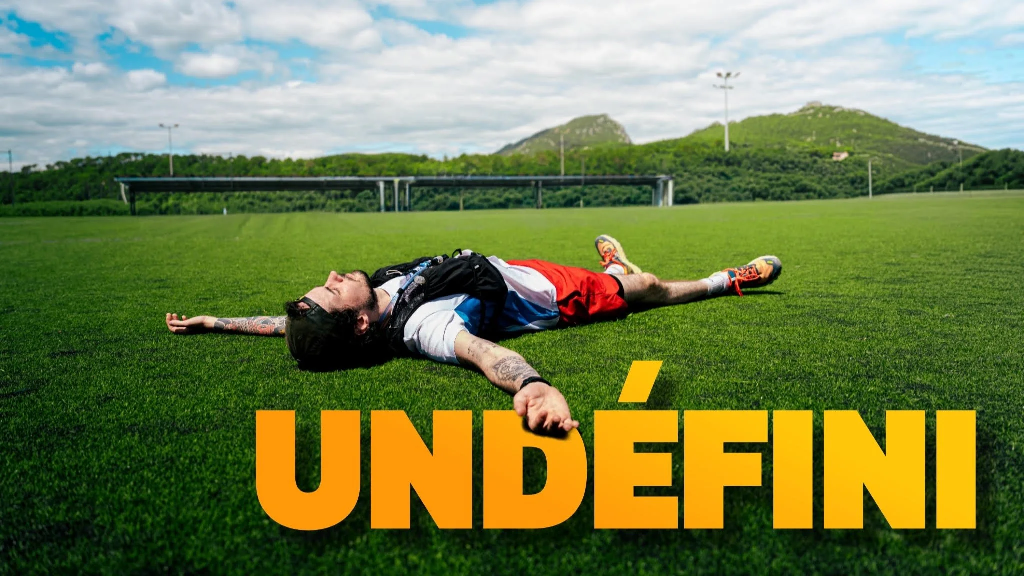 A man laying flat on a green grass field with arms and legs spread out, wearing athletic clothing and a cap, with a landscape of hills and a cloudy sky in the background.