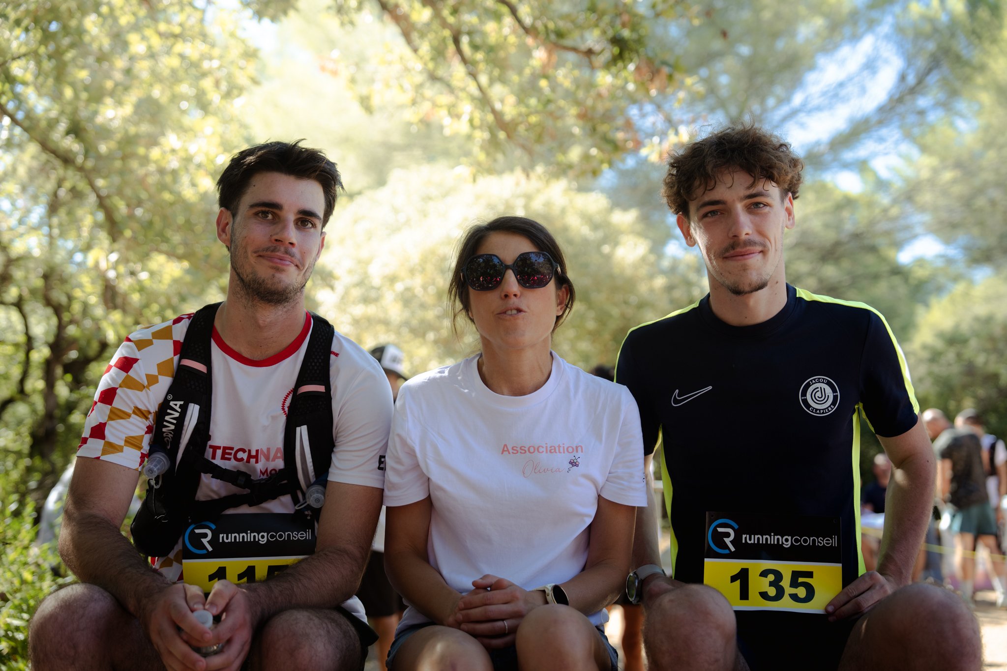 Trois jeunes personnes assises lors d'une course ou d'un événement sportif en plein air, avec des arbres en arrière-plan. Deux hommes et une femme, portant des vêtements de sport, une femme porte des lunettes de soleil et a un t-shirt blanc avec la m