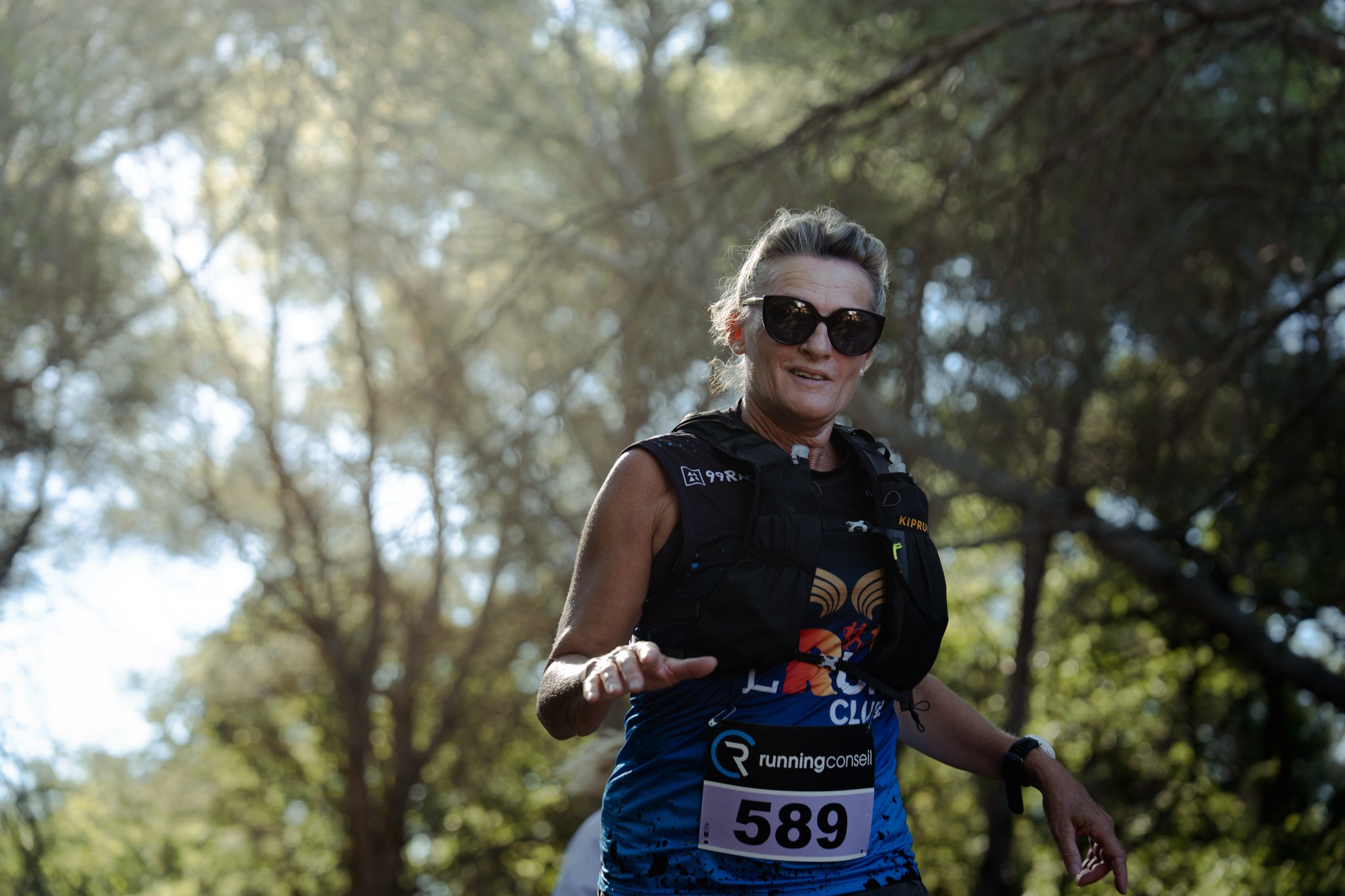 Une femme âgée portant des lunettes noires, un gilet de course, un dossard numéro 589, participe à une course dans une forêt ensoleillée.