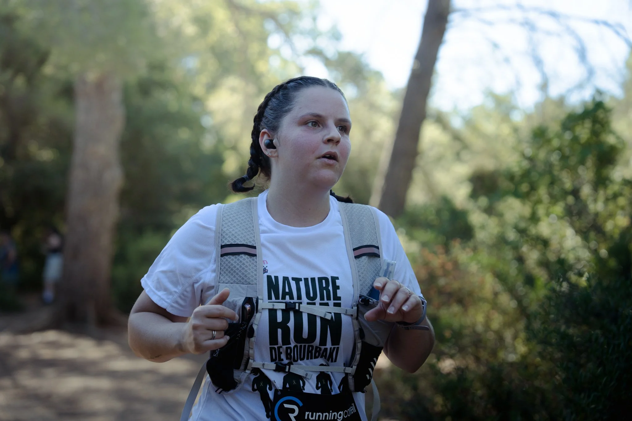 Une femme court dans un parc boisé avec un t-shirt blanc et un sac à dos, portant une oreillette, lors d'une course ou d'un entraînement. Arbres verts en arrière-plan.