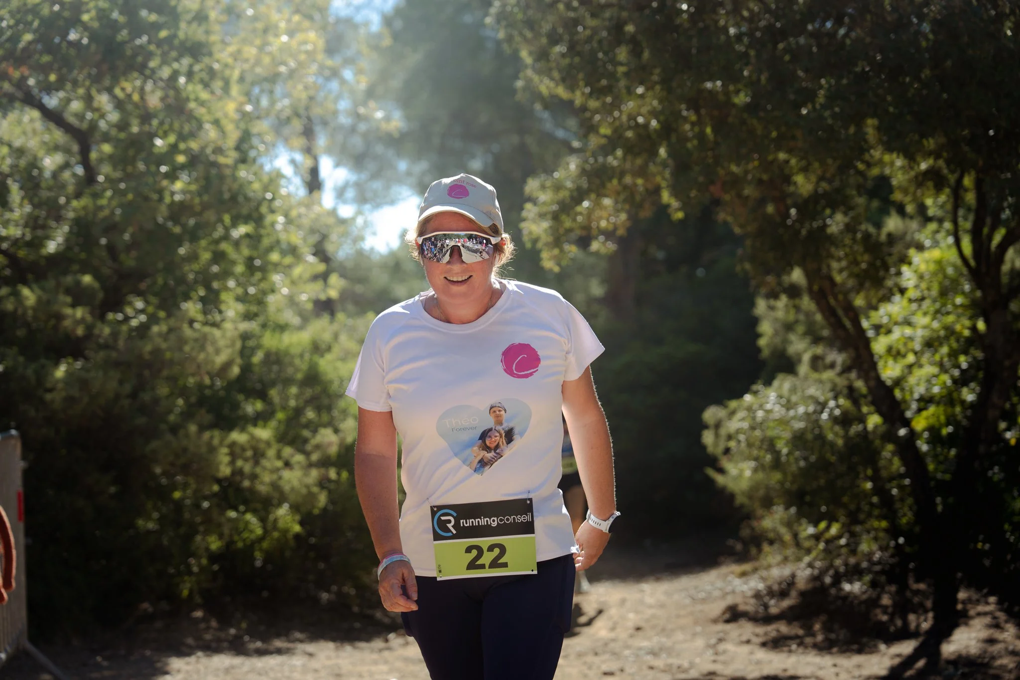 Une femme participe à une course dans un parc arboré le matin, portant une casquette, des lunettes de soleil et un T-shirt avec une photo d'elle-même et d'une autre personne. Elle a un dossard avec le numéro 22.