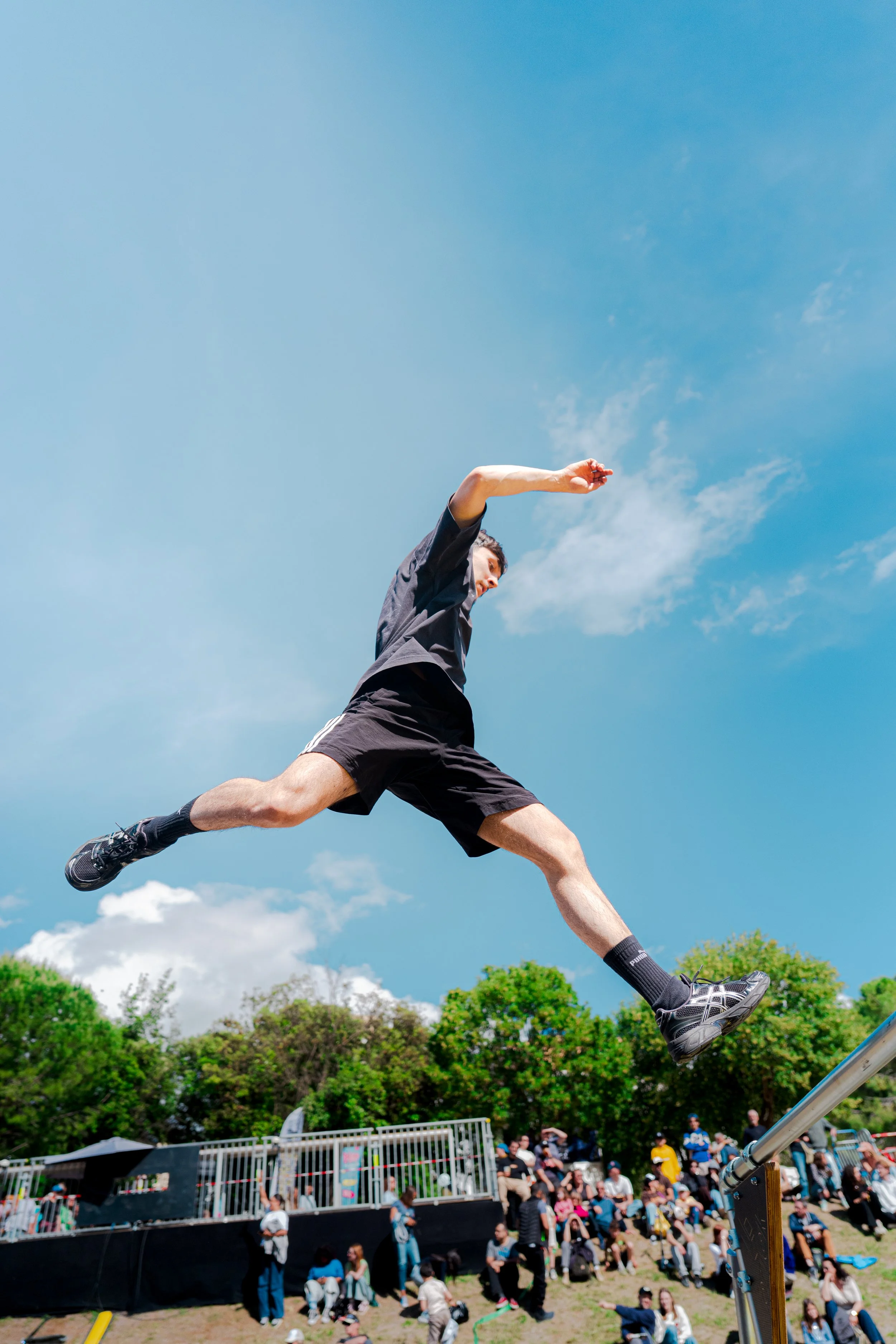 Un homme effectuant un saut acrobatique lors d'une compétition de saut en longueur, avec un public et des arbres en arrière-plan sous un ciel bleu