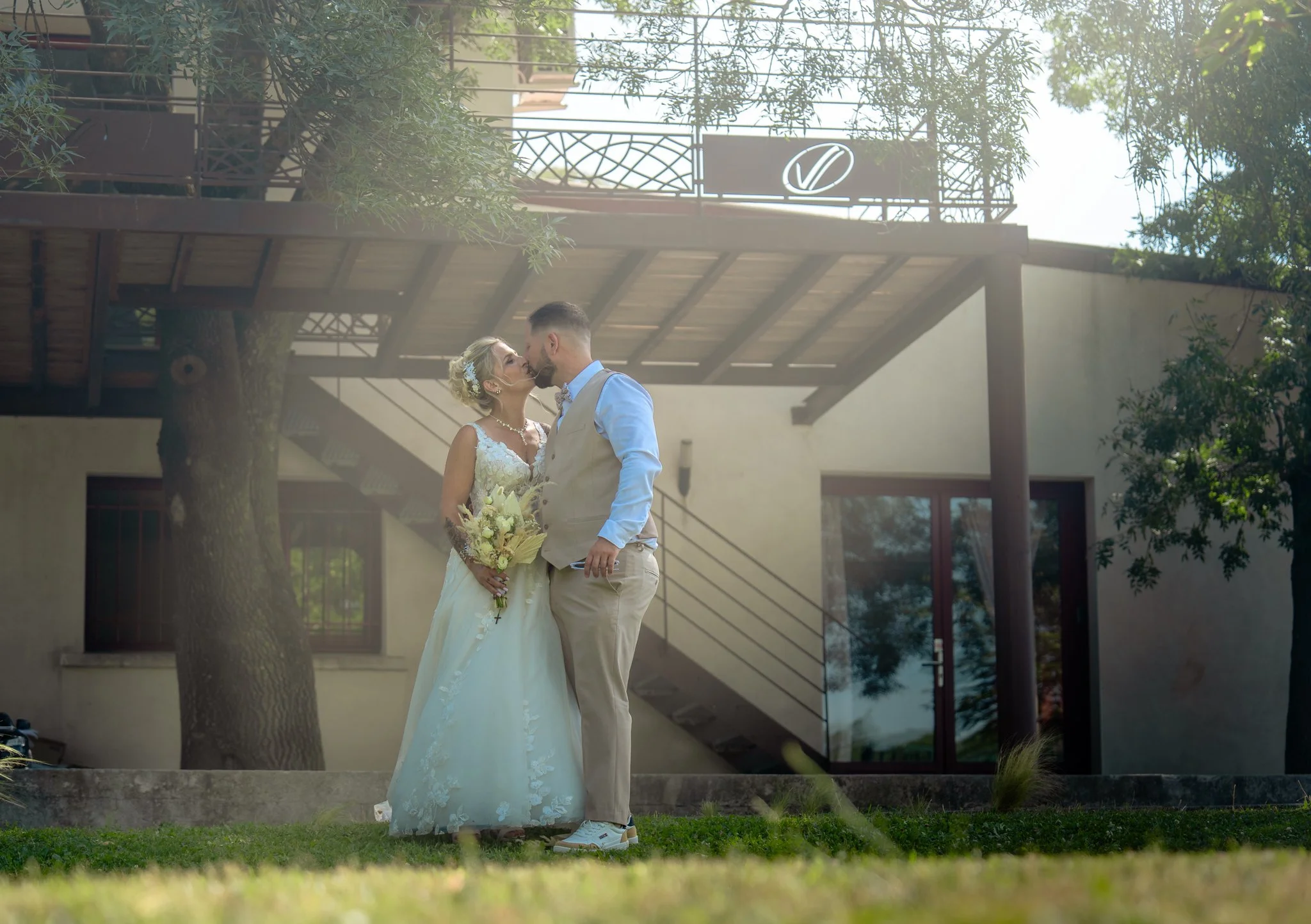 Un couple de mariés s'embrasse devant une maison moderne, entourés d'arbres et de végétation.