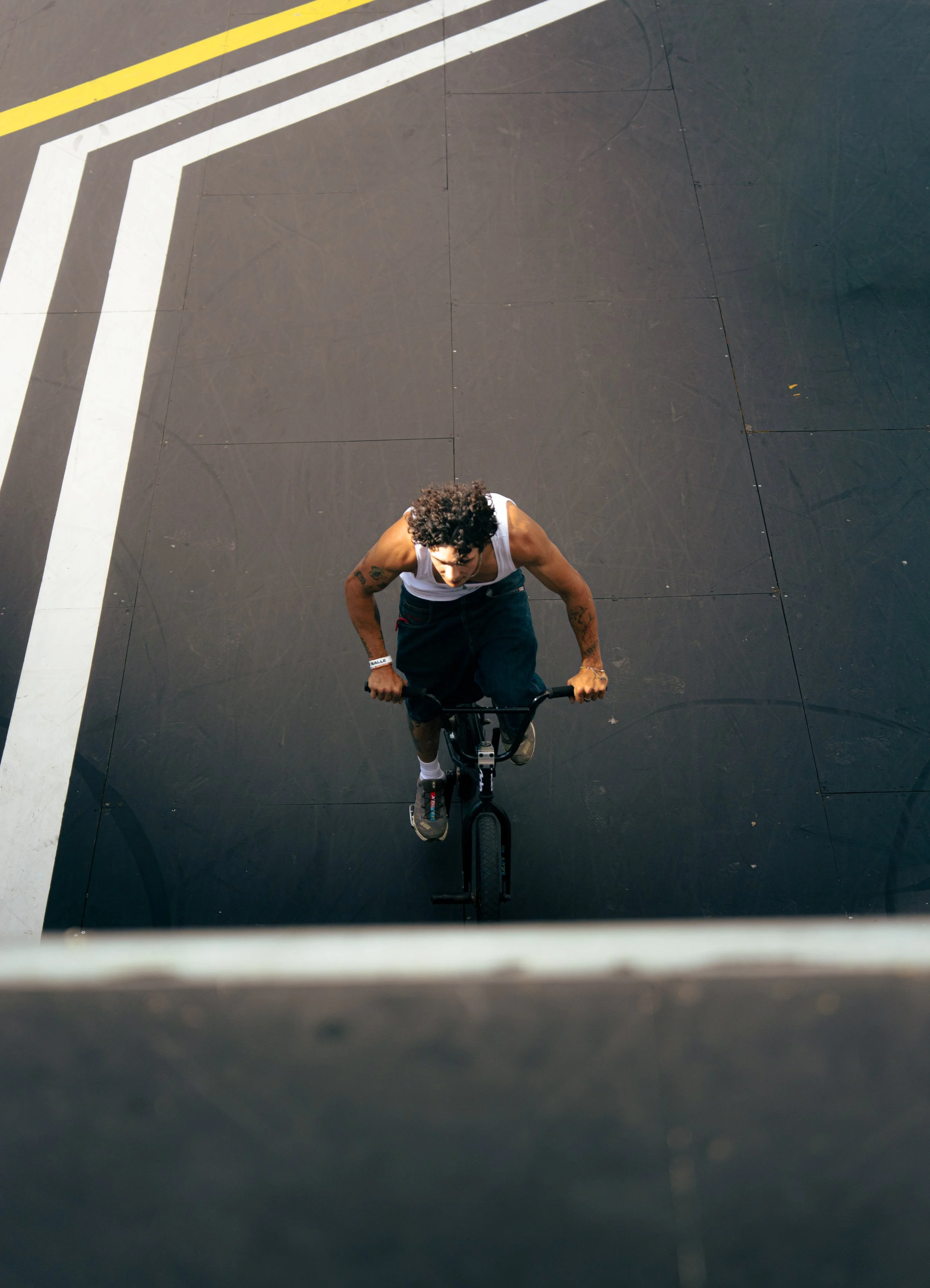 Une personne avec des tatouages, portant un débardeur blanc, faisant du vélo sur une surface noire avec des lignes jaunes et blanches.