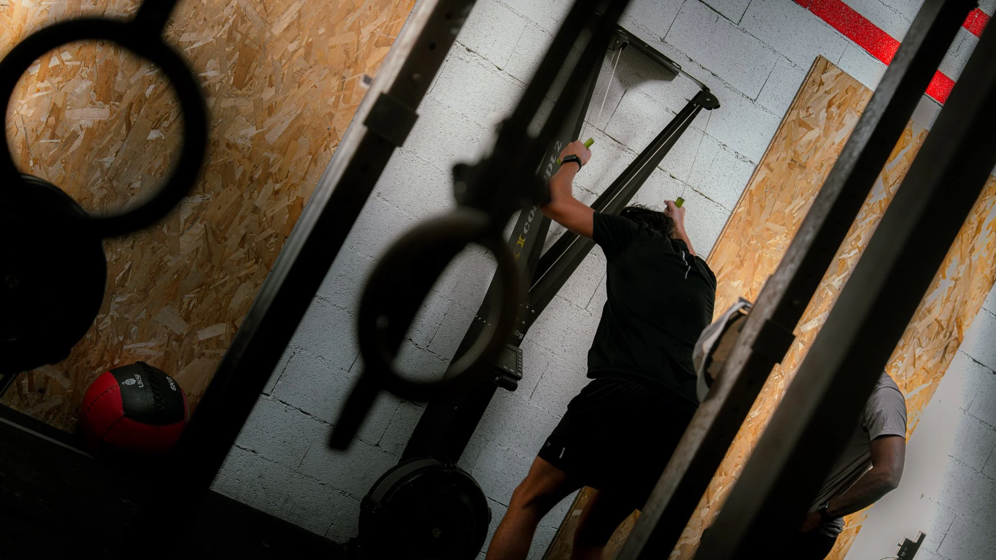 Une personne s'entraînant à la salle de sport, utilisant un appareil d'entraînement de musculation, avec un ballon de médecine posé au sol et un mur en bois derrière elle.