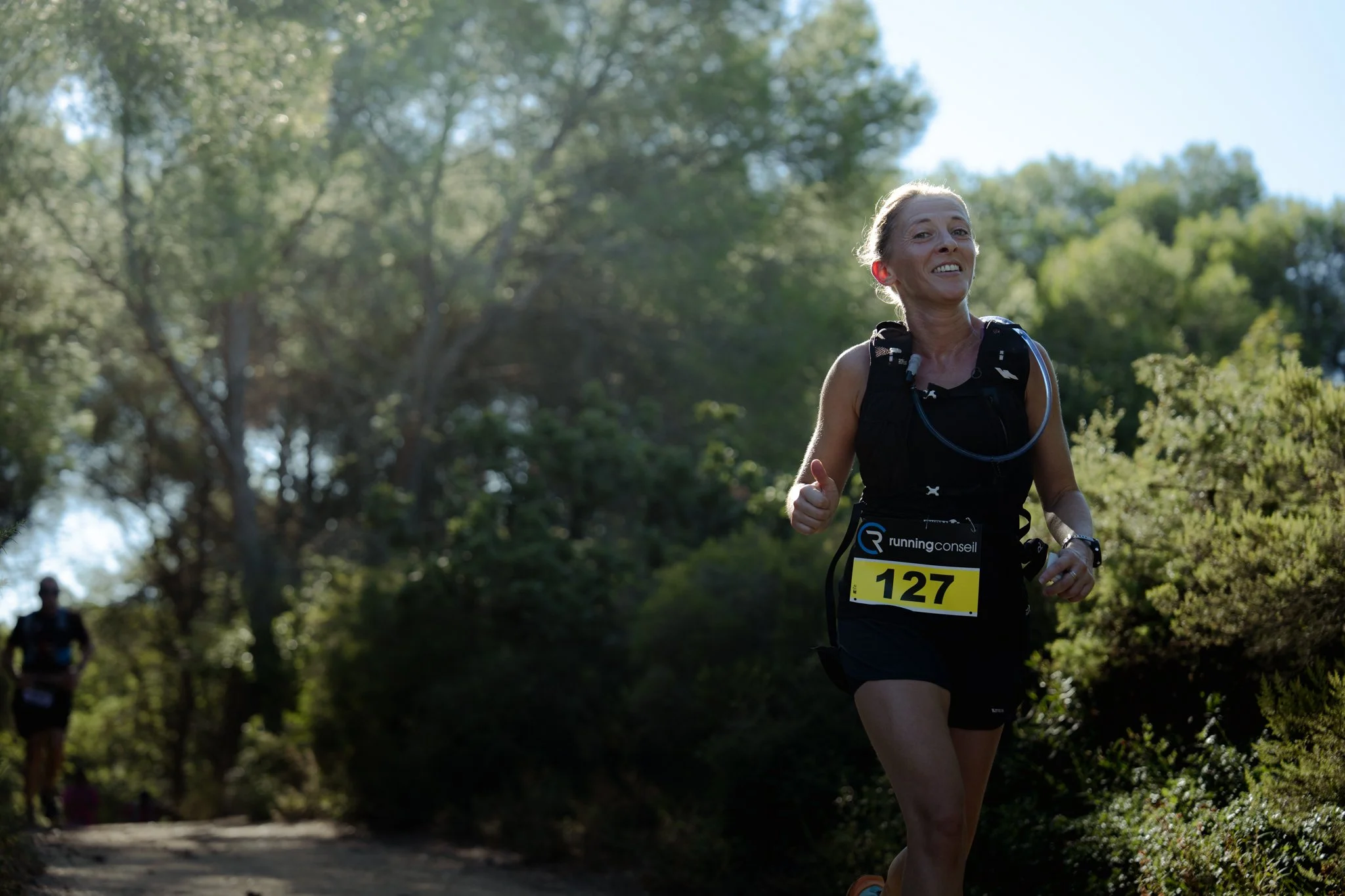 Une femme âgée faisant une course à pied dans un parc ensoleillé, portant un maillot noir, un numéro de course 127, avec un sourire sur le visage.