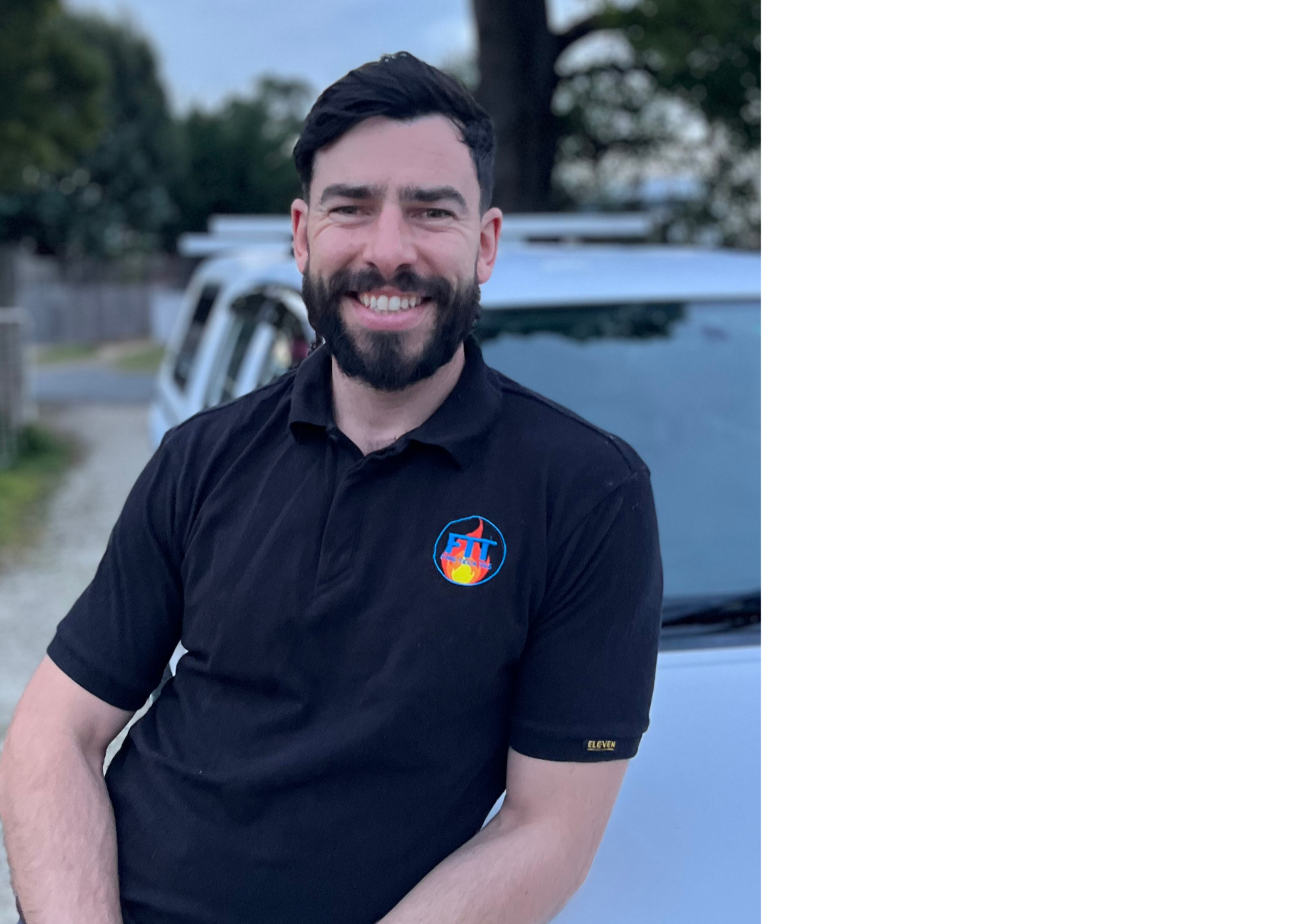 A smiling man with dark hair and a beard, wearing a black shirt with a colorful logo on the chest, standing outdoors with trees and a vehicle in the background.