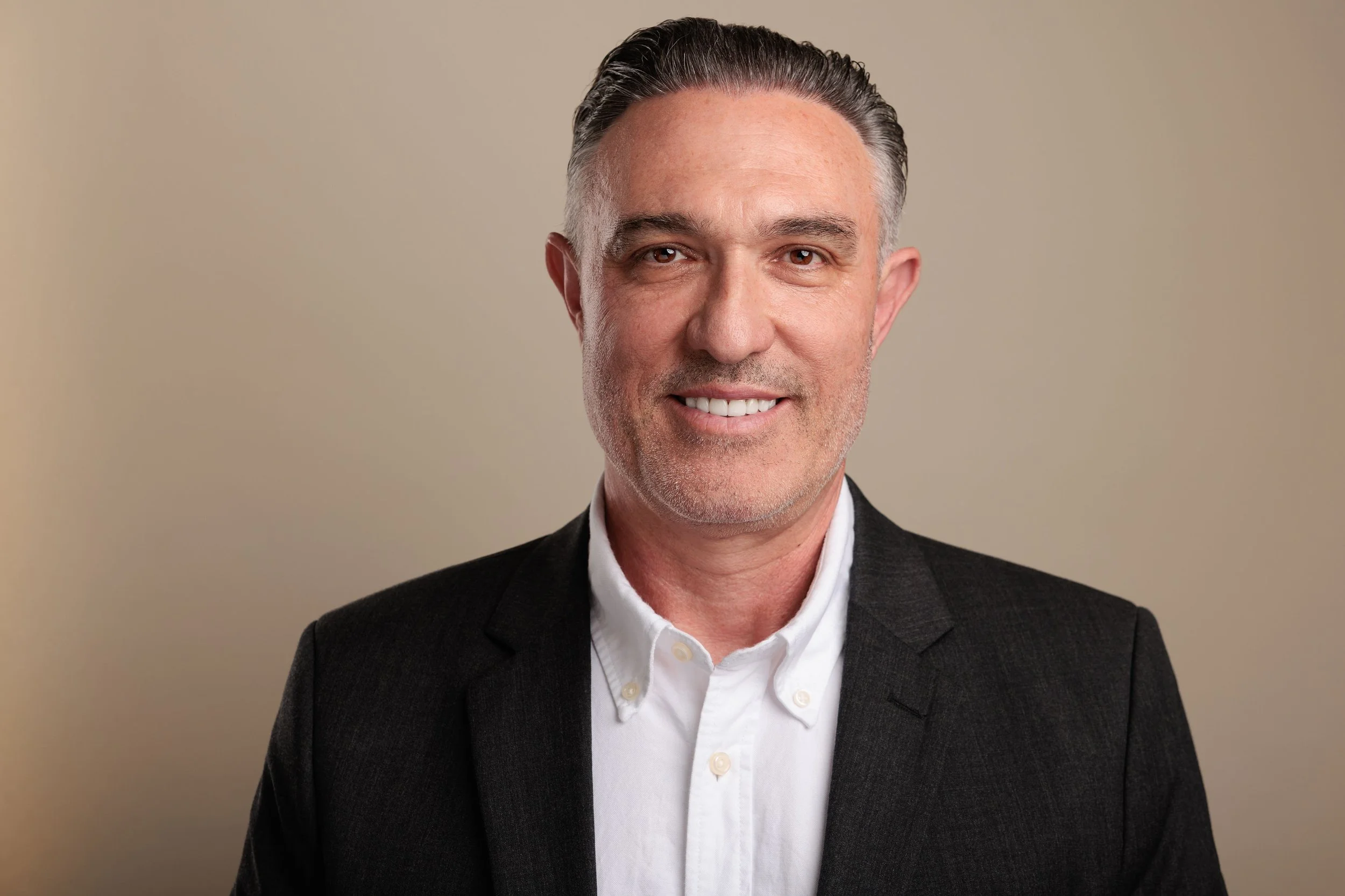 A middle-aged man with short gray hair and brown eyes, smiling, wearing a white collared shirt and a black blazer, against a beige background.