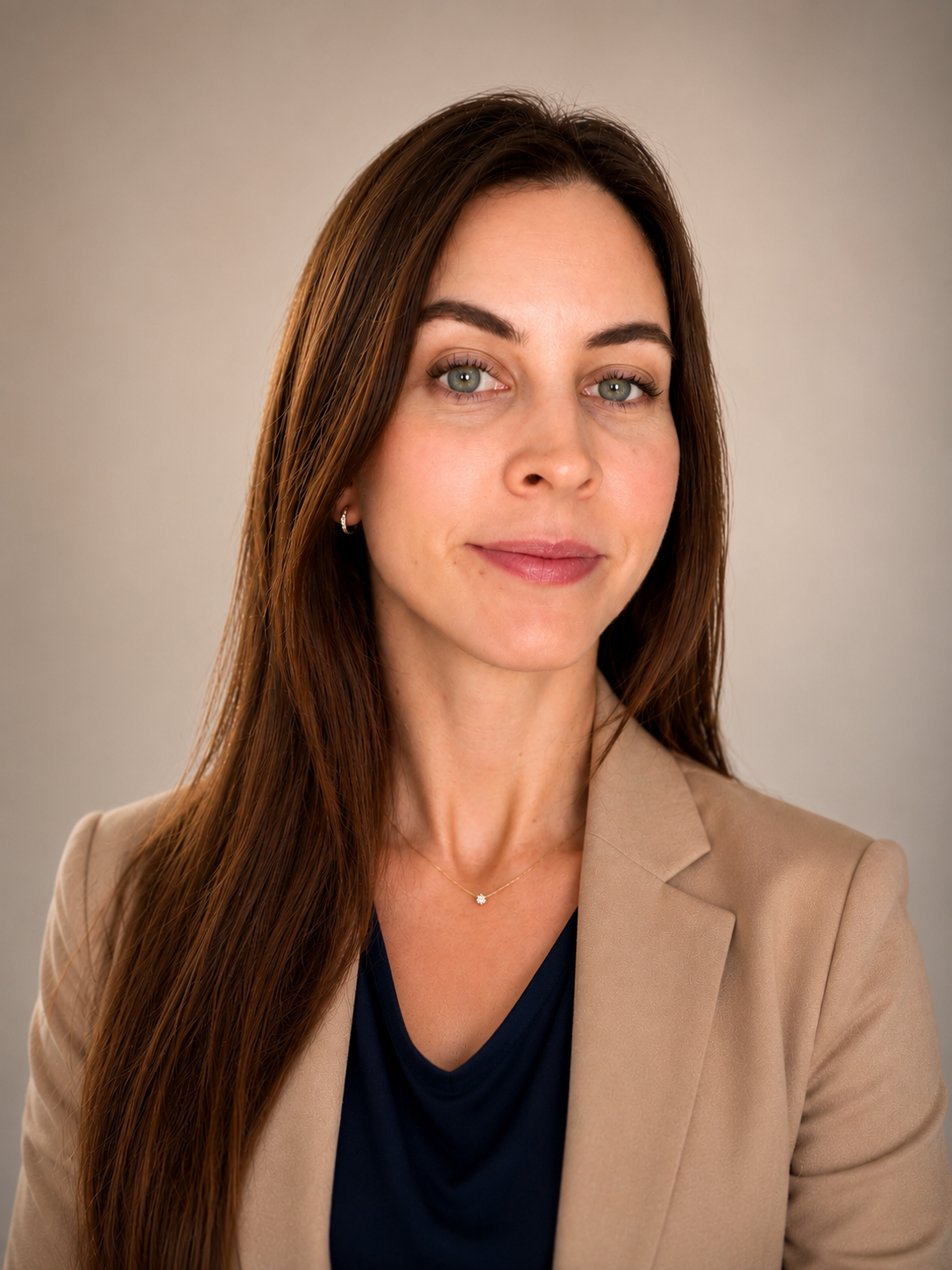 A woman with long brown hair, wearing a beige blazer over a navy top, and a small necklace, facing slightly to the right with a neutral expression against a plain gray background.