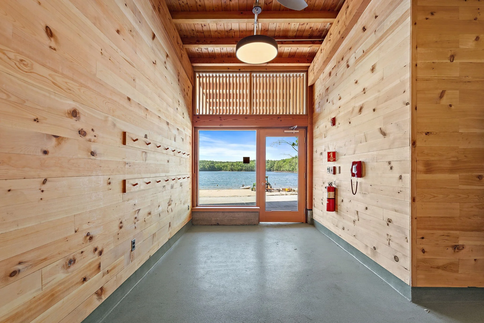 Interior view of a wooden hallway leading to a door with a window, overlooking a lake and tree-lined shoreline under a blue sky.