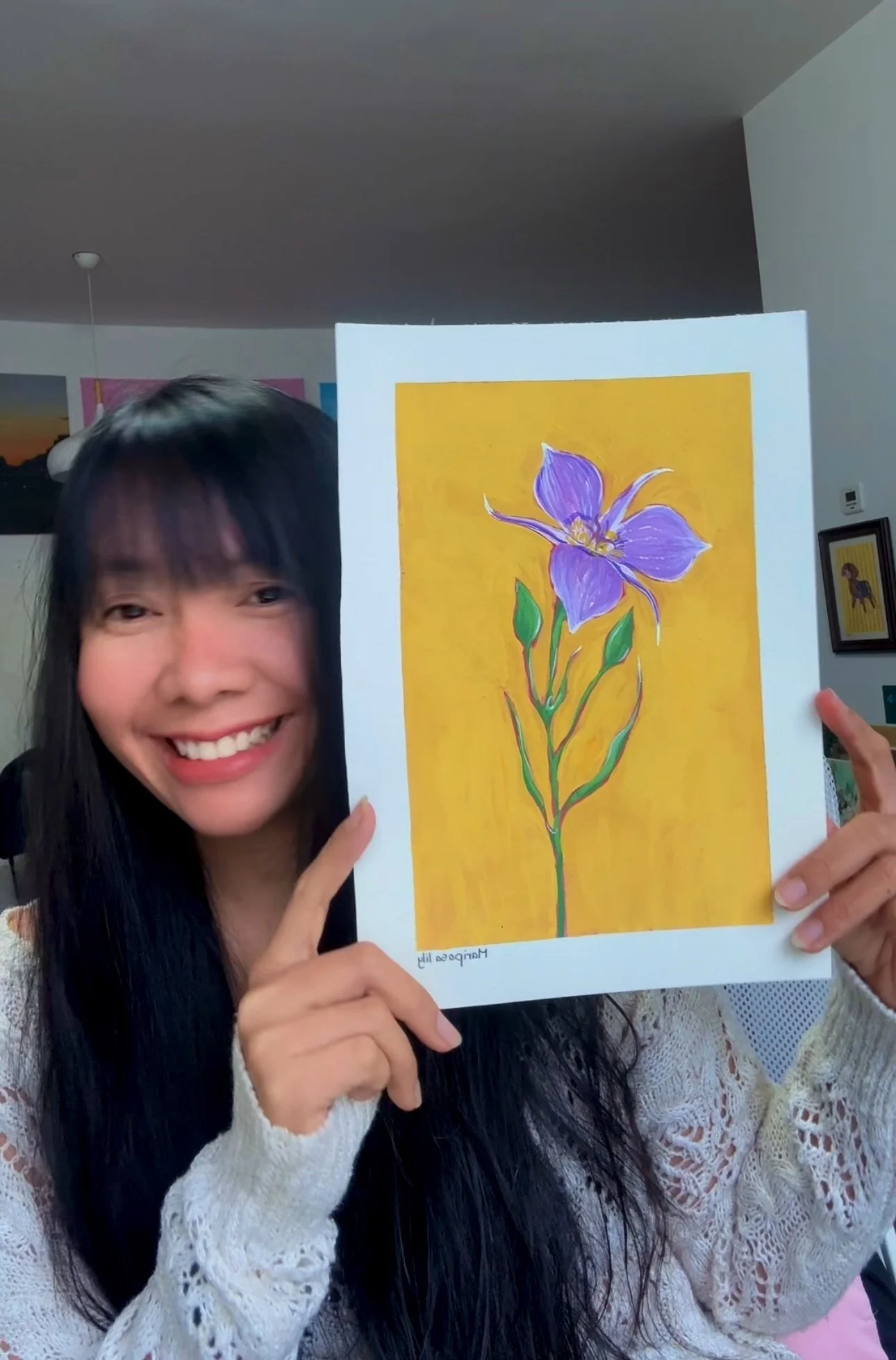 Woman with long black hair smiling, holding a colorful painting of a purple flower with green leaves on a yellow background, in an indoor setting.