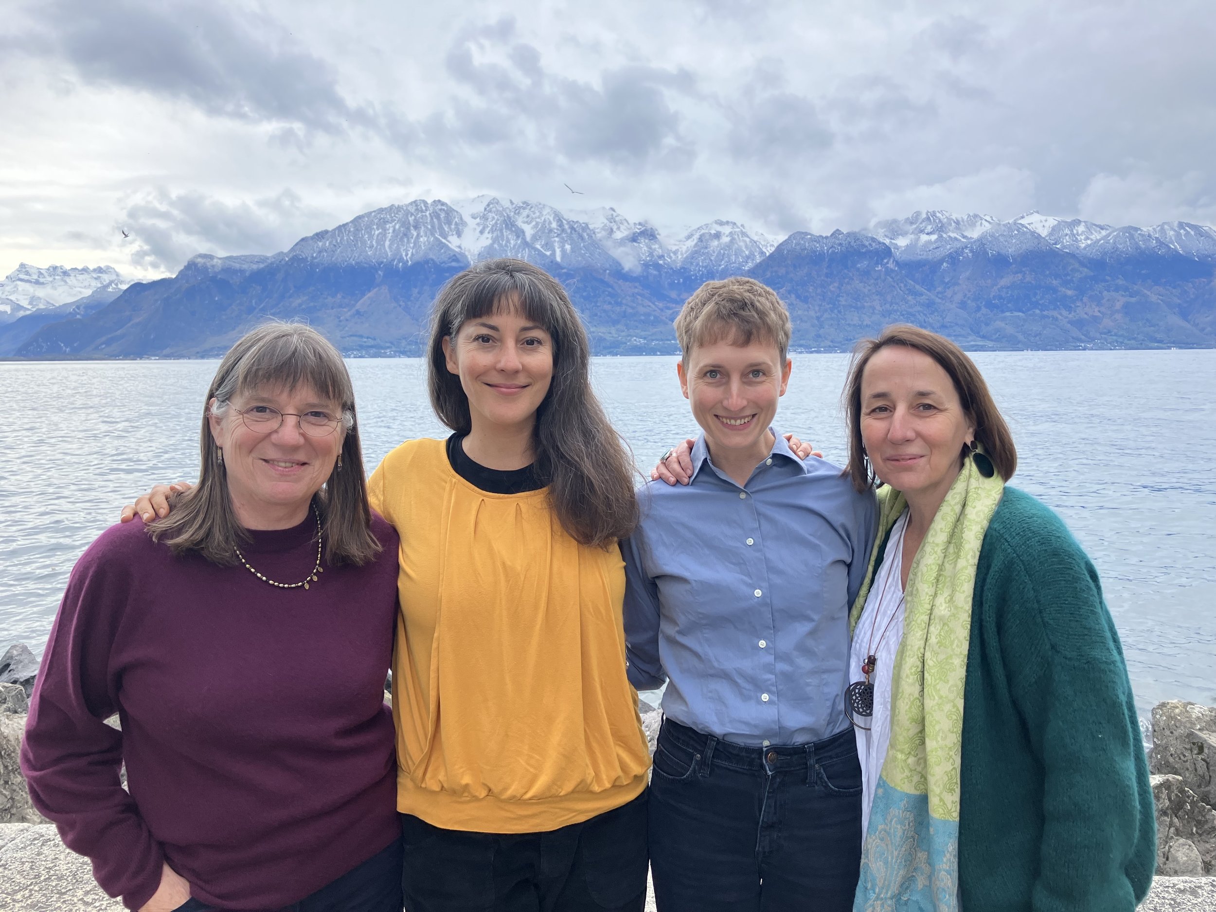 Les membres du collectif souriantes posant près d'un lac avec des montagnes enneigées en arrière-plan, par une journée nuageuse.