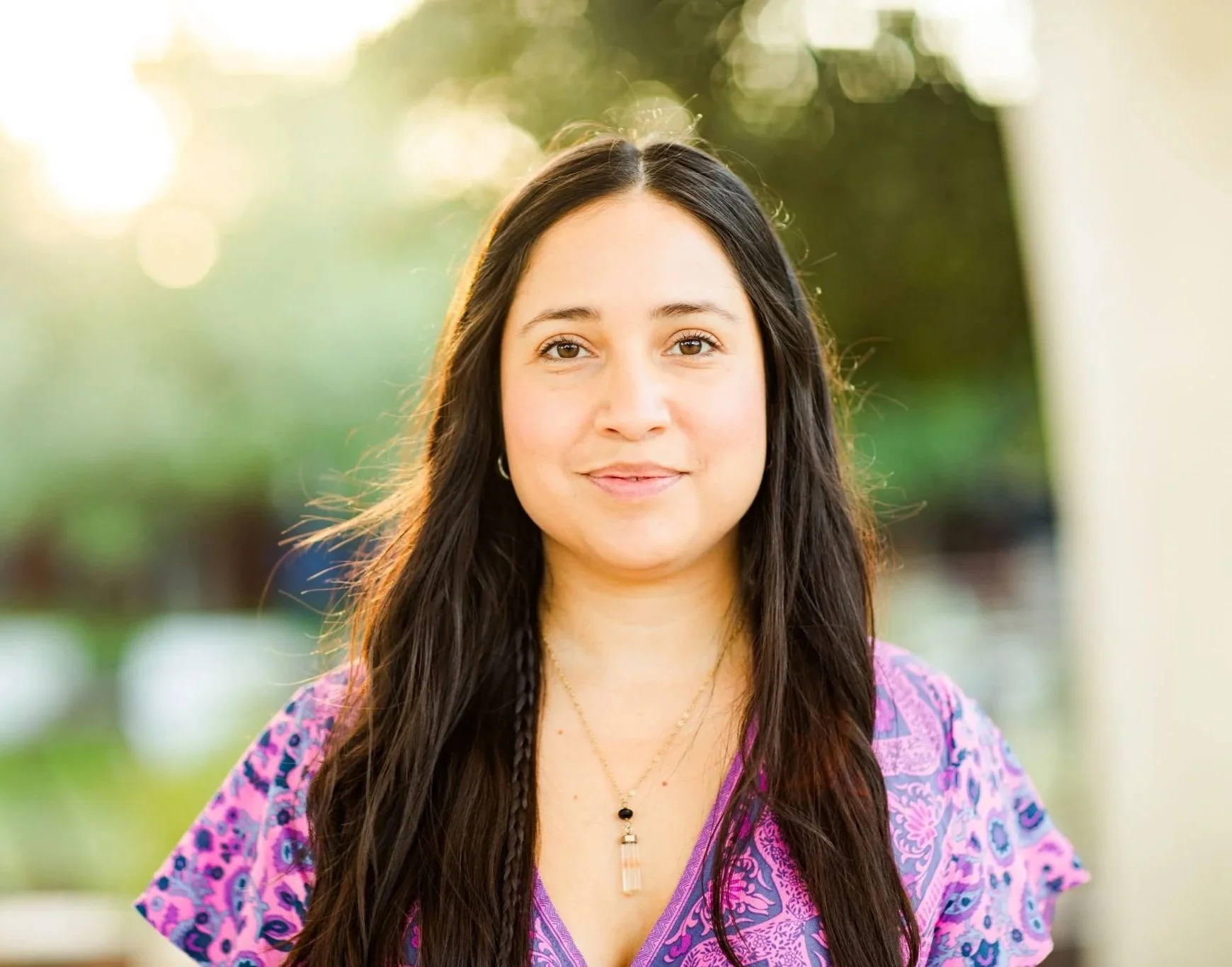 Holistic Latina Counselor in San Antonio, TX
A young woman with long dark hair and light skin, wearing a purple patterned blouse and a necklace, standing outdoors with sunlight and green foliage in the background.