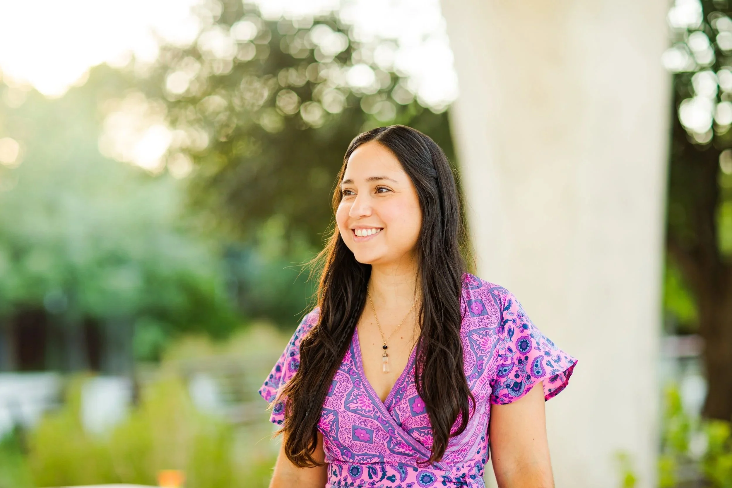 Telehealth Therapy in Texas
A smiling woman with long dark hair wearing a purple and pink patterned dress standing outdoors near water and greenery.