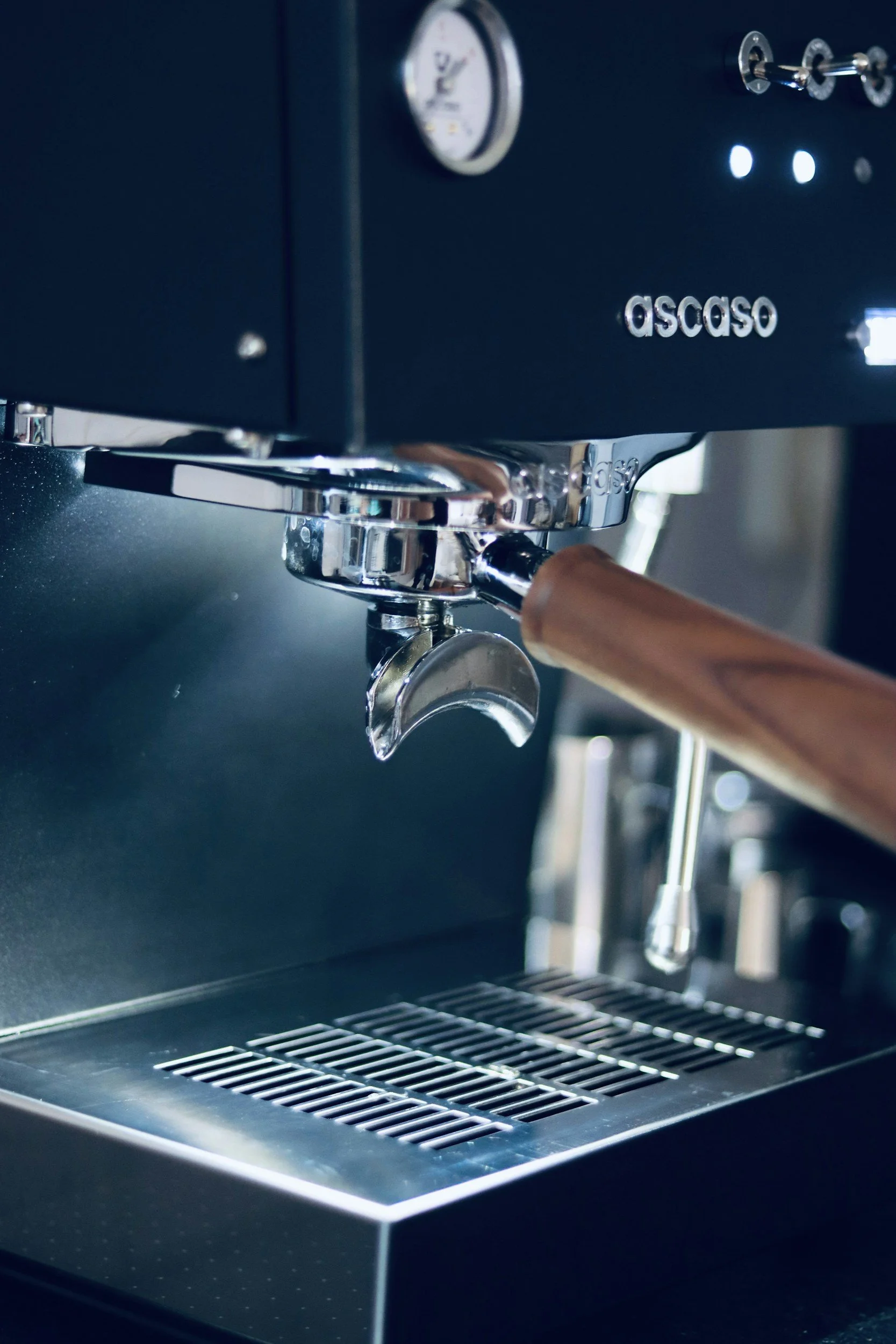 Nahaufnahme einer Espressomaschine mit Holzgriff, Logo auf der Maschine, Metallteilen und Rahmen in Schwarz, im Hintergrund Tassenhalter.