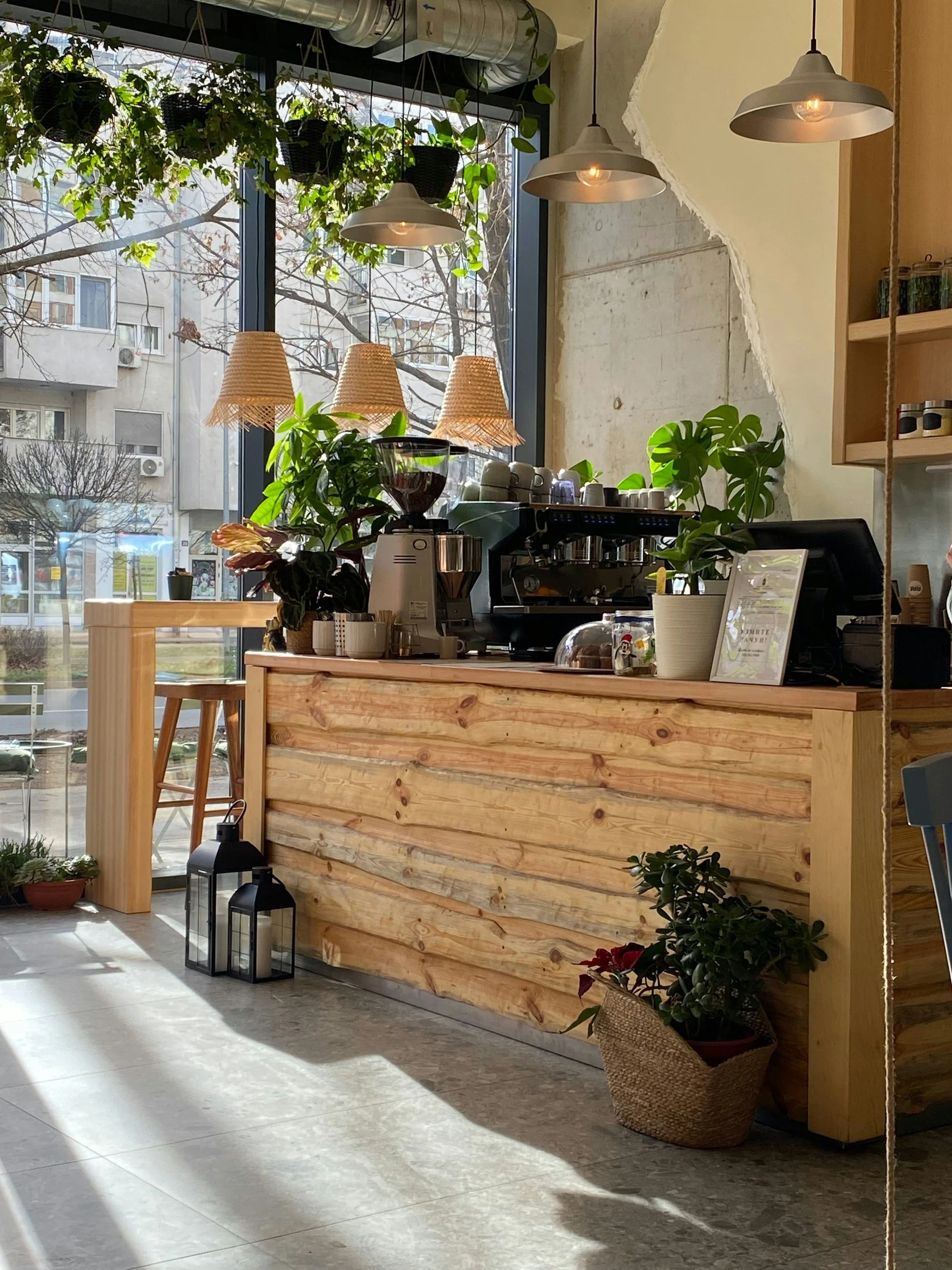Innenaufnahme eines Café oder einer Bar mit viel Holzdekoration, Pflanzen und großem Fenster. Es hängen Lampen von der Decke, im Hintergrund steht eine Kaffeemaschine auf der Theke, einige Pflanzen und Dekorationsartikel sind sichtbar.