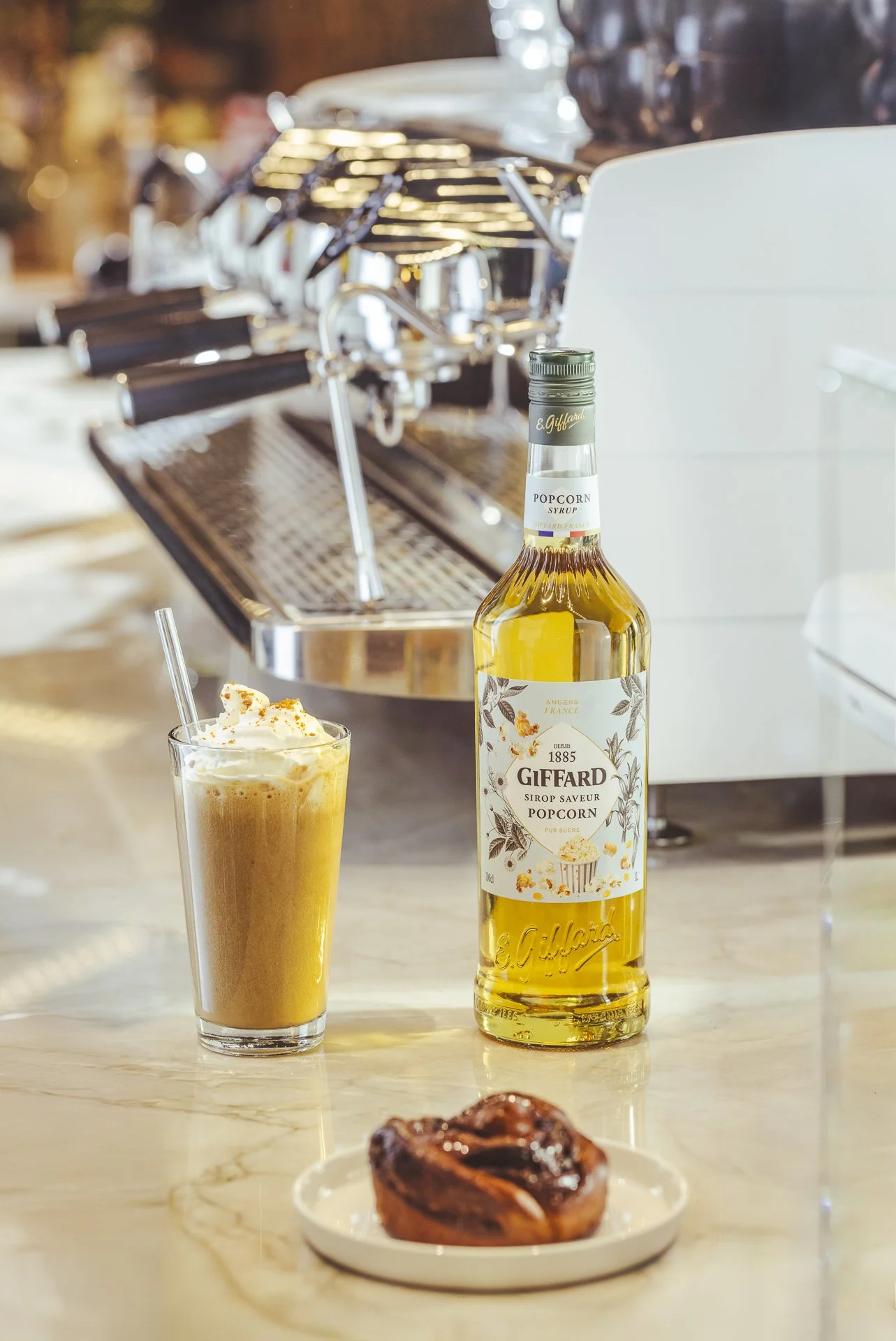 Ein Glas mit einem Cappuccino, eine Flasche Popcorn-Sirup und ein Schokodonut auf einem weißen Teller auf einer Marmor-Tischplatte, im Hintergrund eine Kaffeemaschine.