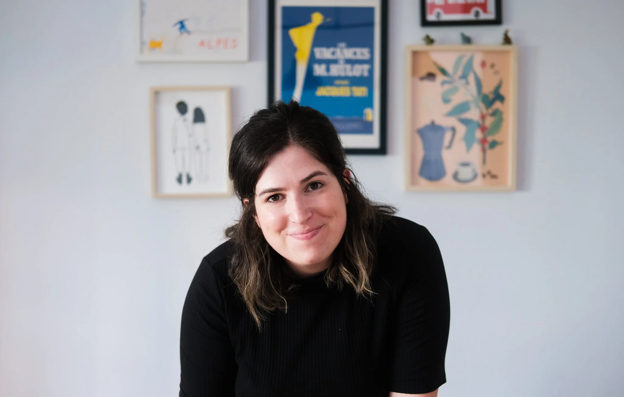 A woman with dark brown hair wearing a black top, smiling, in front of a white wall with framed artwork and posters.