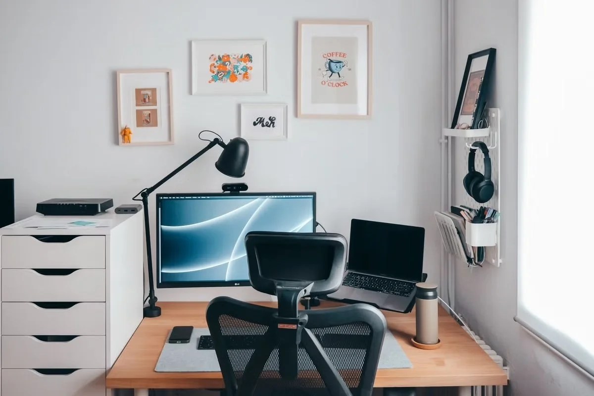 Home office workspace with a wooden desk, computer monitor, laptop, black ergonomic chair, desk lamp, and wall-mounted shelving with headphones, framed pictures, and artwork. White drawers on the left and decorative framed art on the white wall.