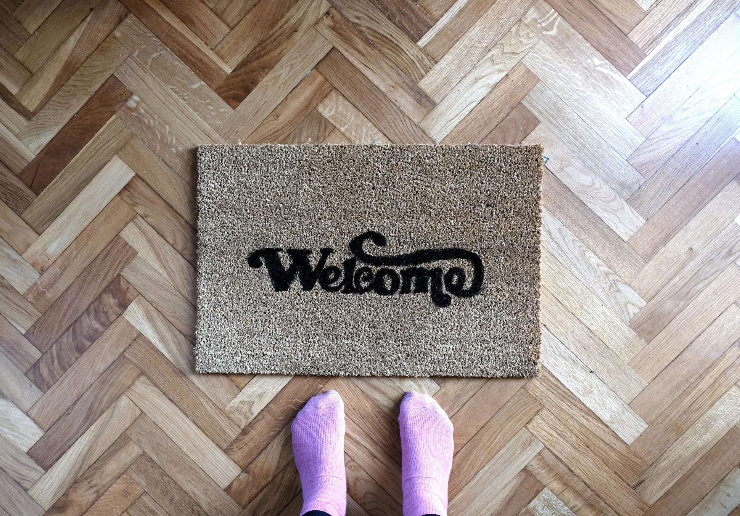 Top-down view of a doormat on a hardwood floor with the word 'Welcome' written on it, and a person wearing purple socks standing in front of it.