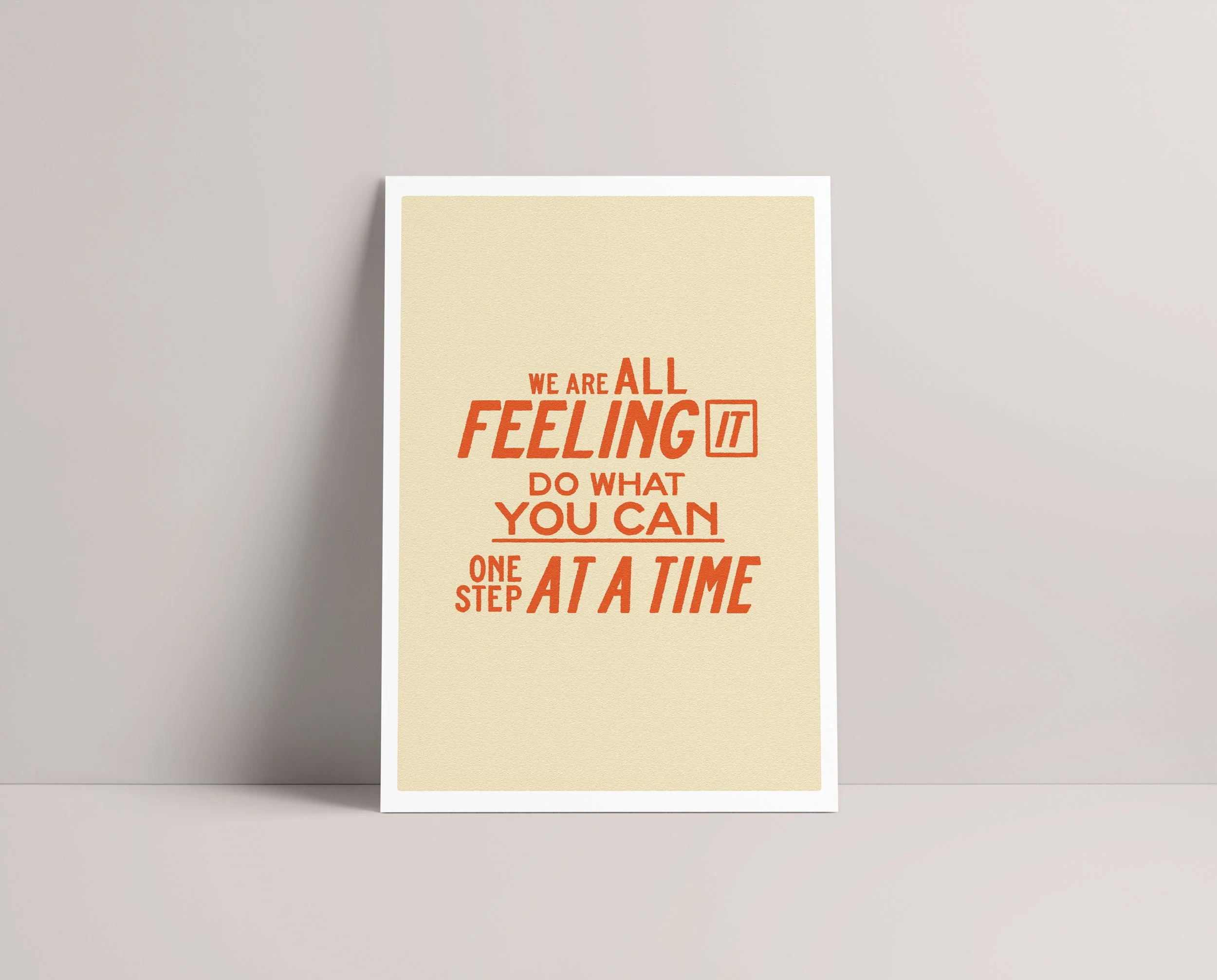 A beige-colored paper with red text reading, 'We are all feeling it do what you can one step at a time,' leaning against a light gray wall.