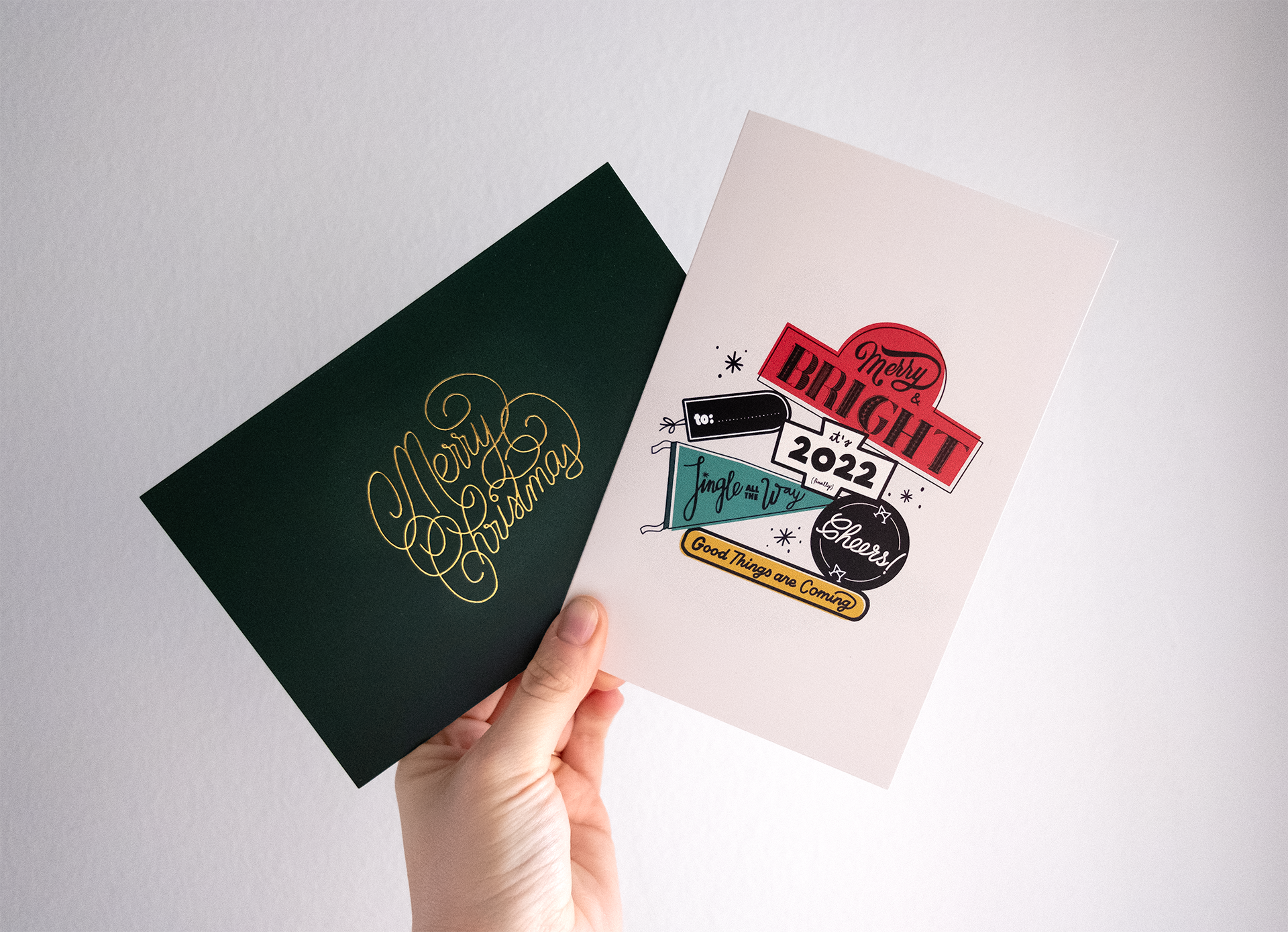 A person holding two Christmas greeting cards: one black with gold script reading "Merry Christmas" and one light pink with colorful vintage-styled graphics and text including "Merry & Bright 2022," "to:," "Jingle all the way," "Cheers!" and "Good th