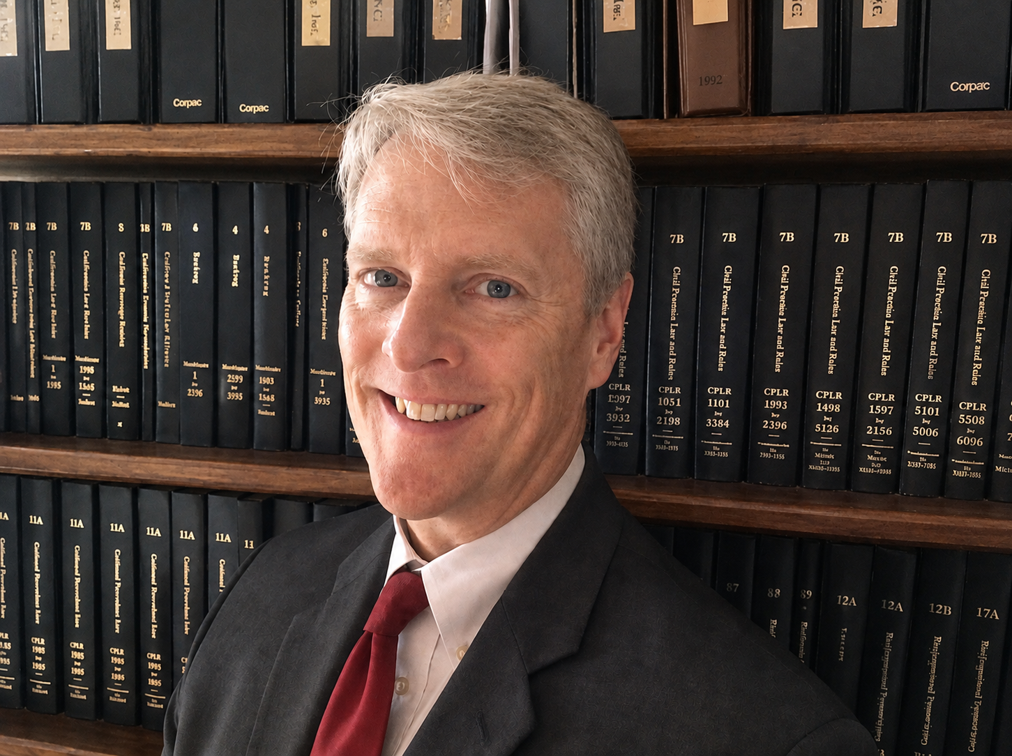A man wearing a suit and red tie, smiling, in front of shelves filled with black books with gold lettering.