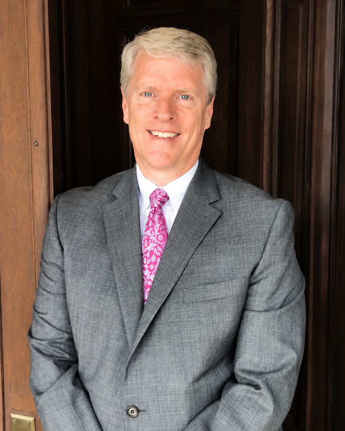 A smiling man in a gray suit and pink patterned tie standing in front of a wooden panel background.