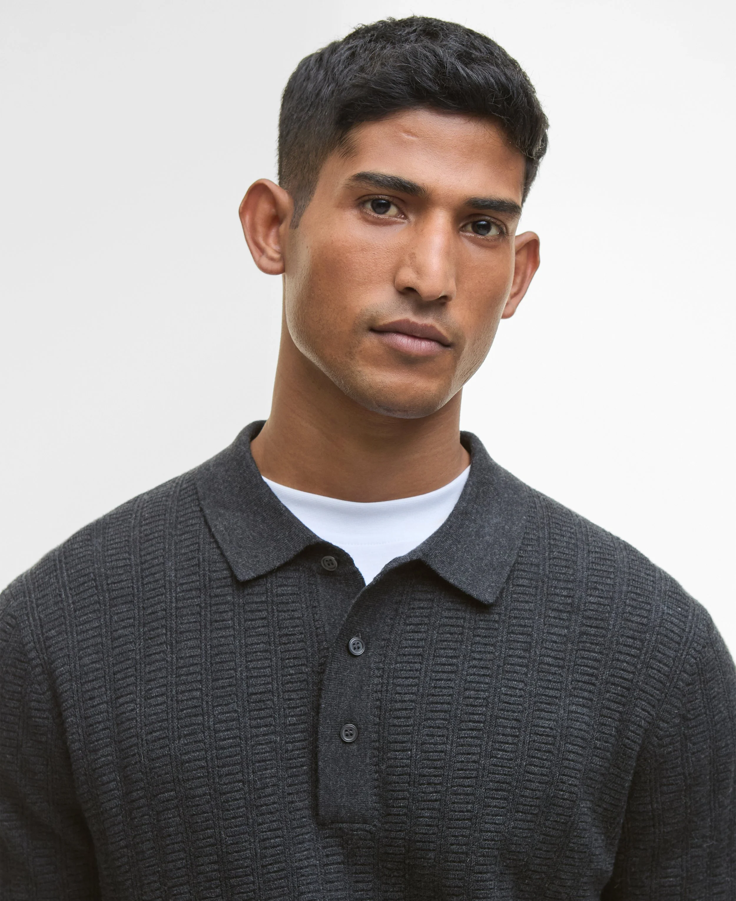 Close-up of a young man with short dark hair, tanned skin, wearing a dark gray ribbed sweater over a white shirt, against a plain white background.