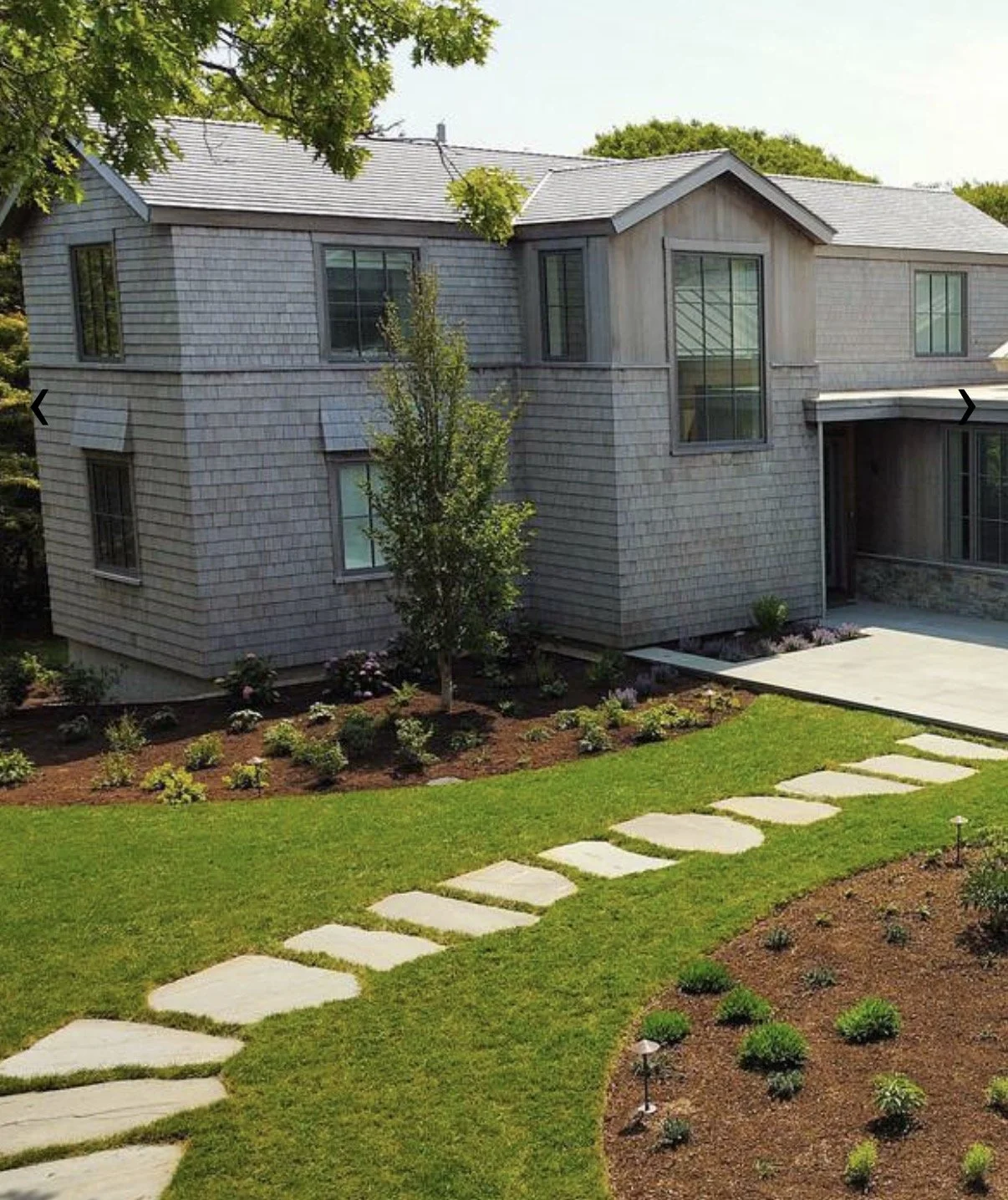 A modern two-story house with gray siding and a sloped roof, surrounded by a landscaped yard with a stone pathway, green grass, small plants, and trees.