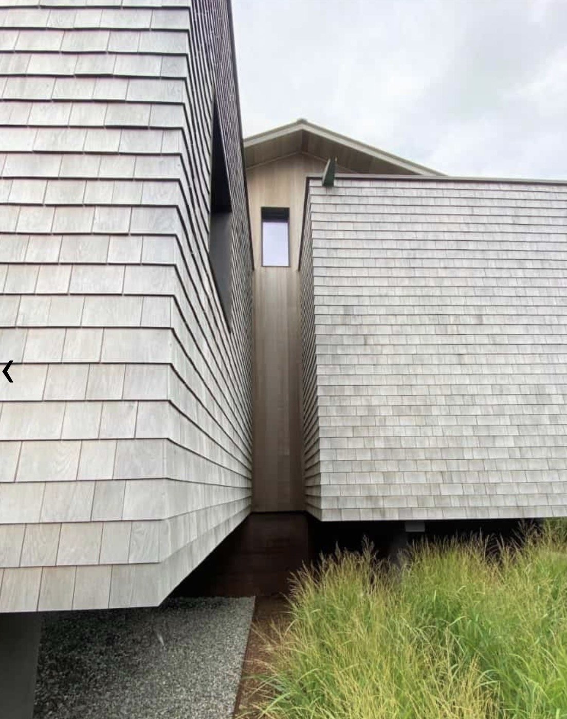 A modern house with shingled exterior walls and narrow space between building sections, with grass in the foreground and cloudy sky overhead.