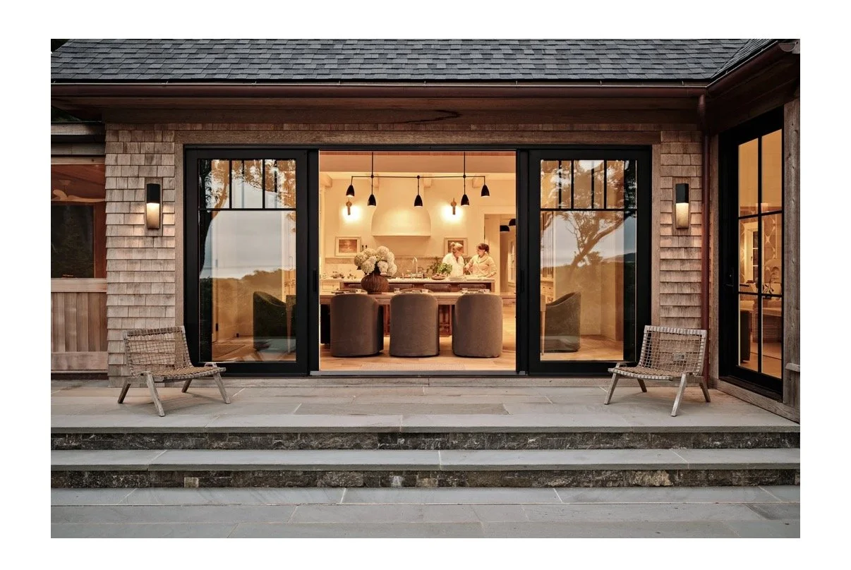View of a modern house with large glass sliding doors opening to a well-lit interior dining room with people inside. The house has a stone and wooden exterior with two outdoor wicker chairs on the patio.