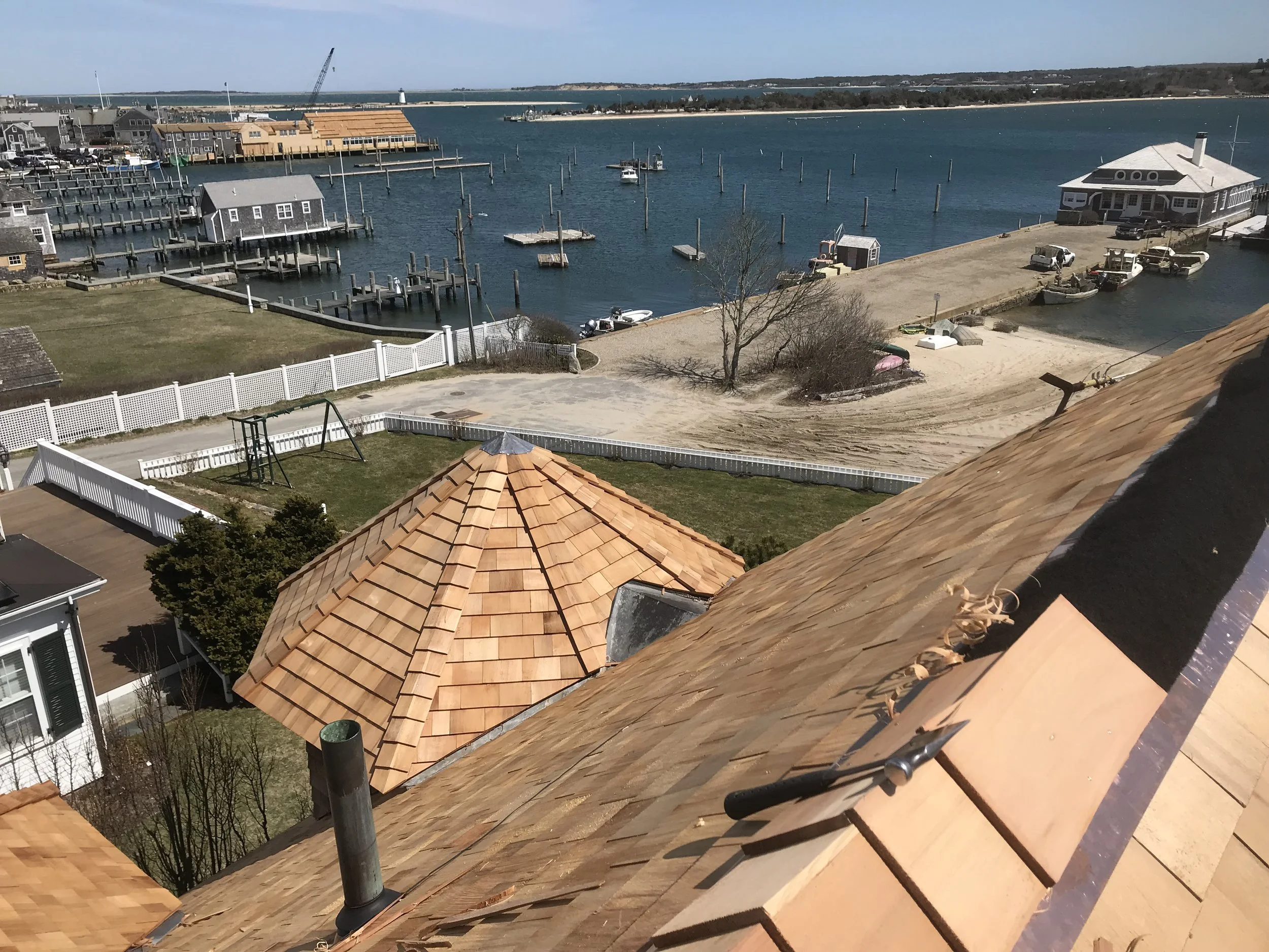 View from a rooftop showing houses with shingled roofs, a fenced yard with a swing set, and a body of water in the background with boats and docks.