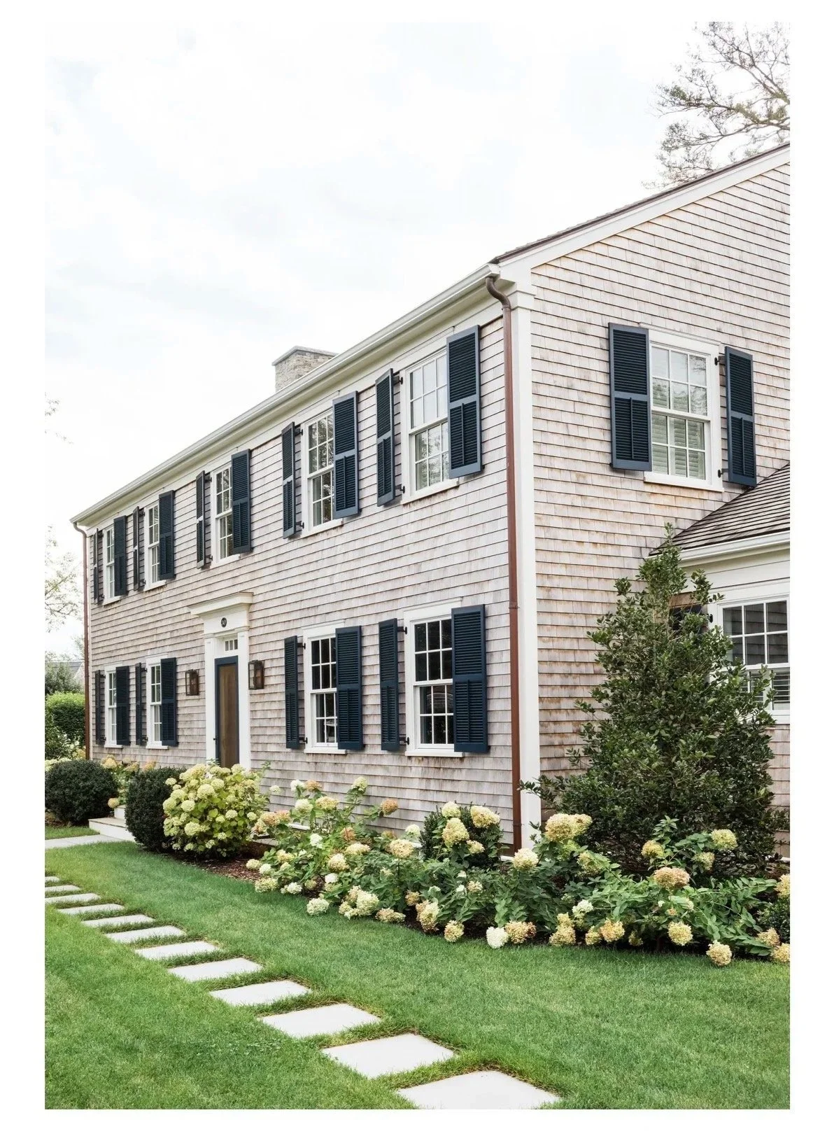 A two-story house with beige wooden clapboard siding, black shutters on multiple windows, a dark front door, and a white decorative pediment over the door. The house is surrounded by a well-maintained garden with lush green lawn, flowering bushes, an