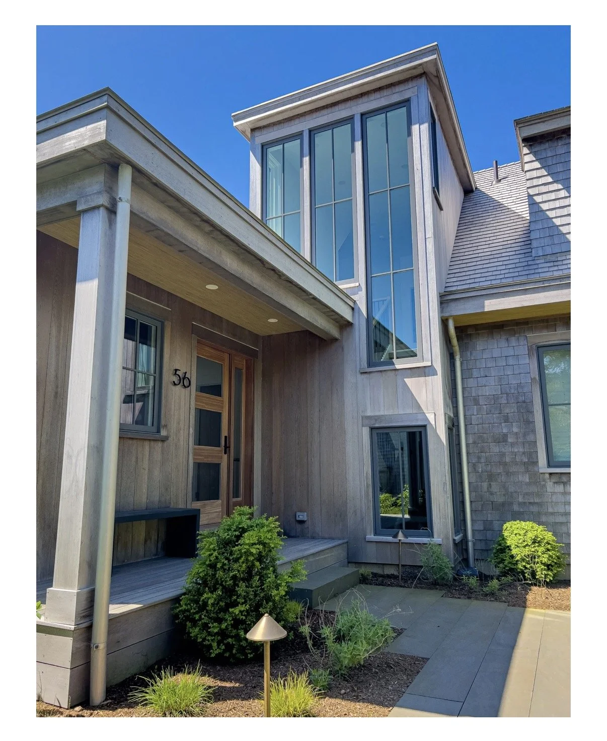 Close-up of a modern house's entrance featuring a wooden door, large glass windows, and a small garden with shrubs and a pathway, under a clear blue sky.