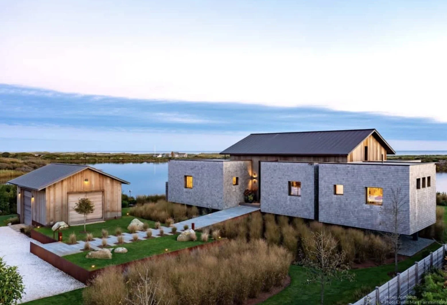Modern house with gray stone and wooden siding, surrounded by a landscaped yard, near water with marshy land in the background, under a partly cloudy sky.