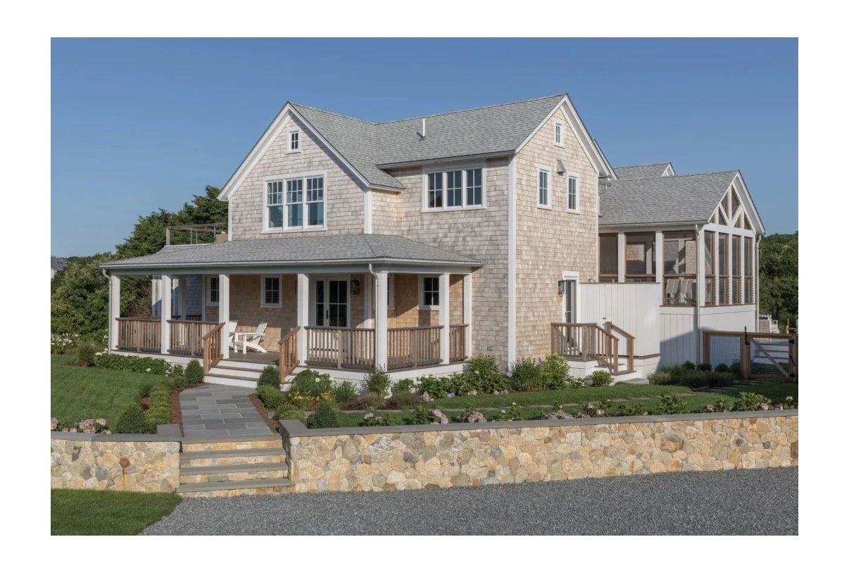 A two-story house with a shingle exterior, front porch with wooden railings and steps, surrounded by a landscaped garden with bushes and flowers, and a stone retaining wall at the front.