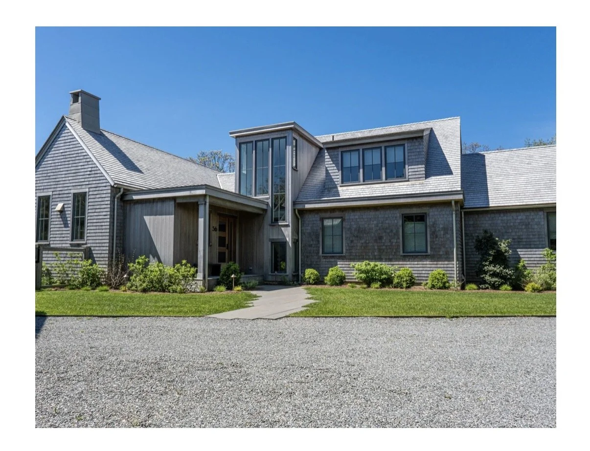 A modern residential house with gray shingle and wood siding, multiple windows, and a small front garden with bushes, on a clear day with blue sky.