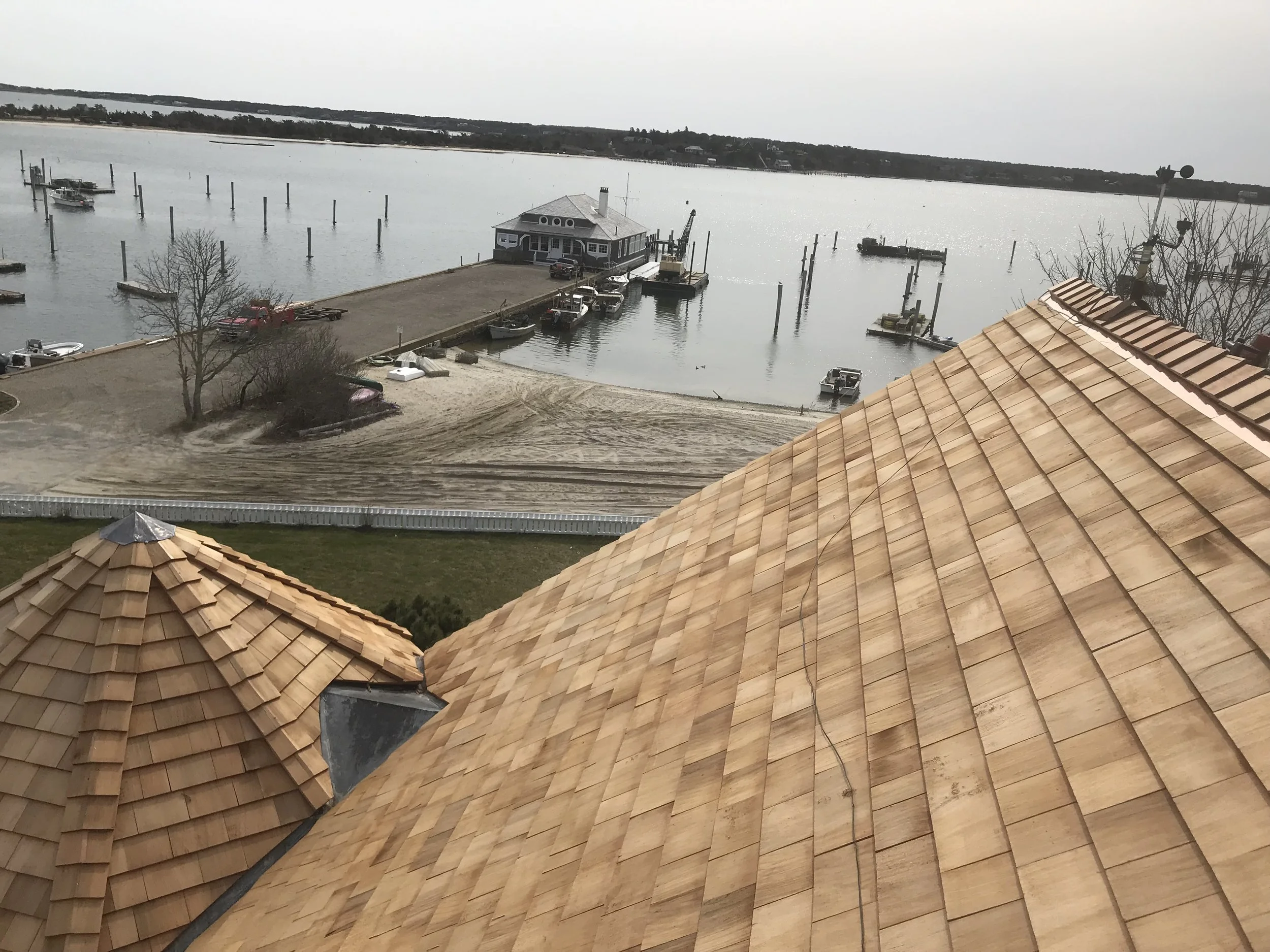 View of a waterfront with boats, a dock, and a house under a cloudy sky, seen from a rooftop with tan shingles.