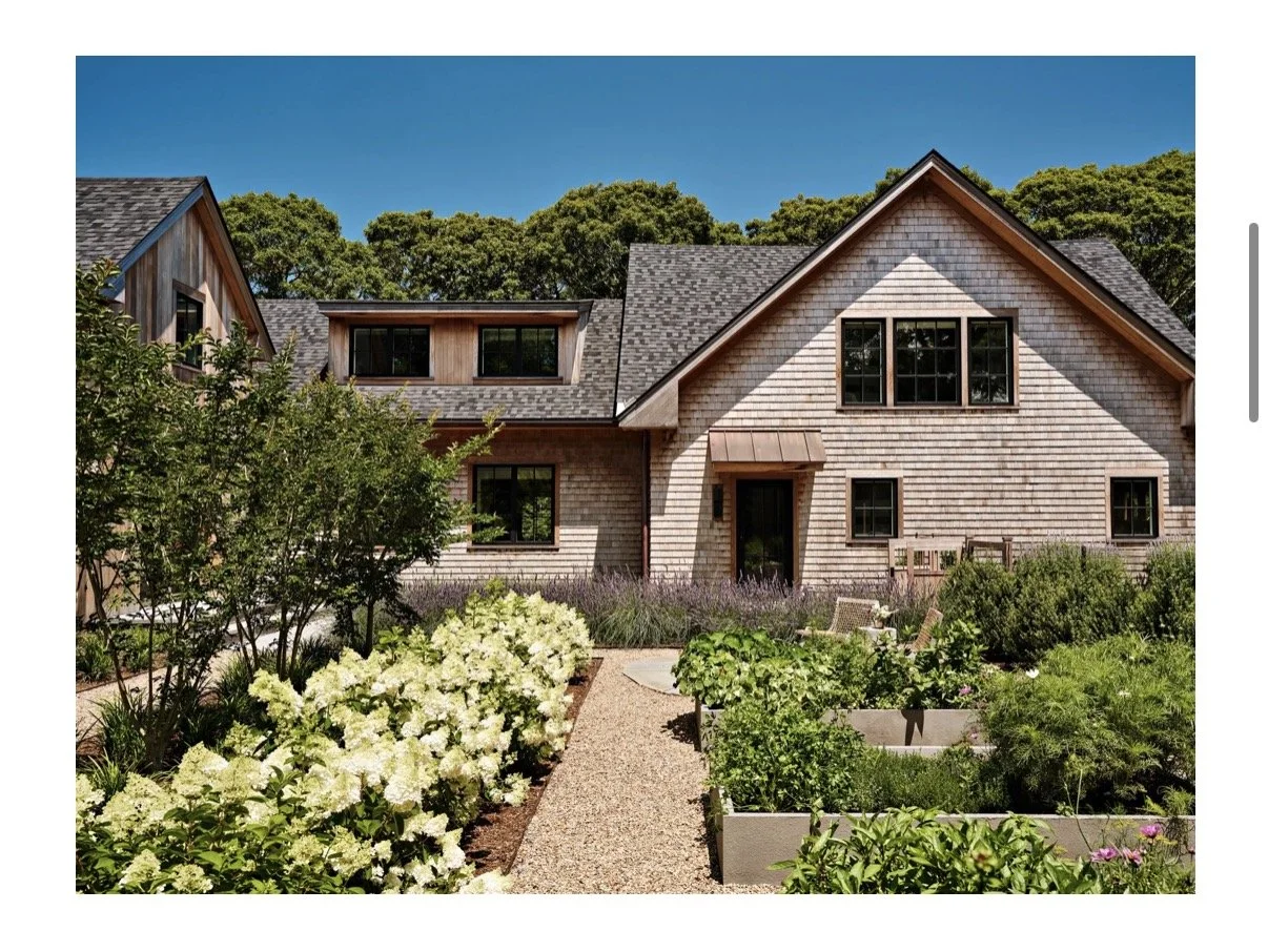 Front view of a modern house with wooden siding, black framed windows, a gabled roof, and a landscaped garden with a gravel walkway and various shrubs and flowers.