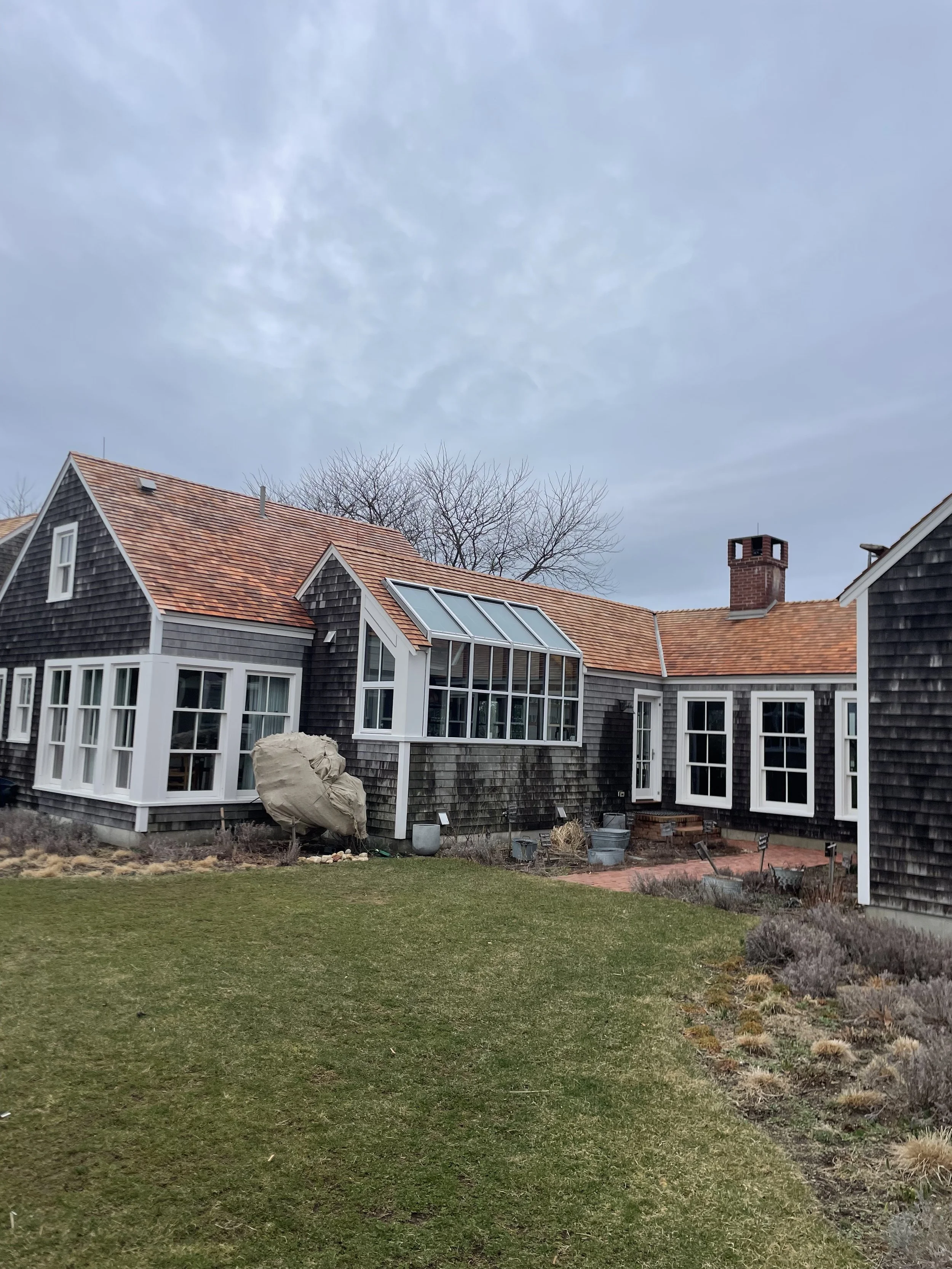 A house with black shingle siding, a reddish-tile roof, and a large glass sunroom, under an overcast sky, with a green lawn and garden in the foreground.