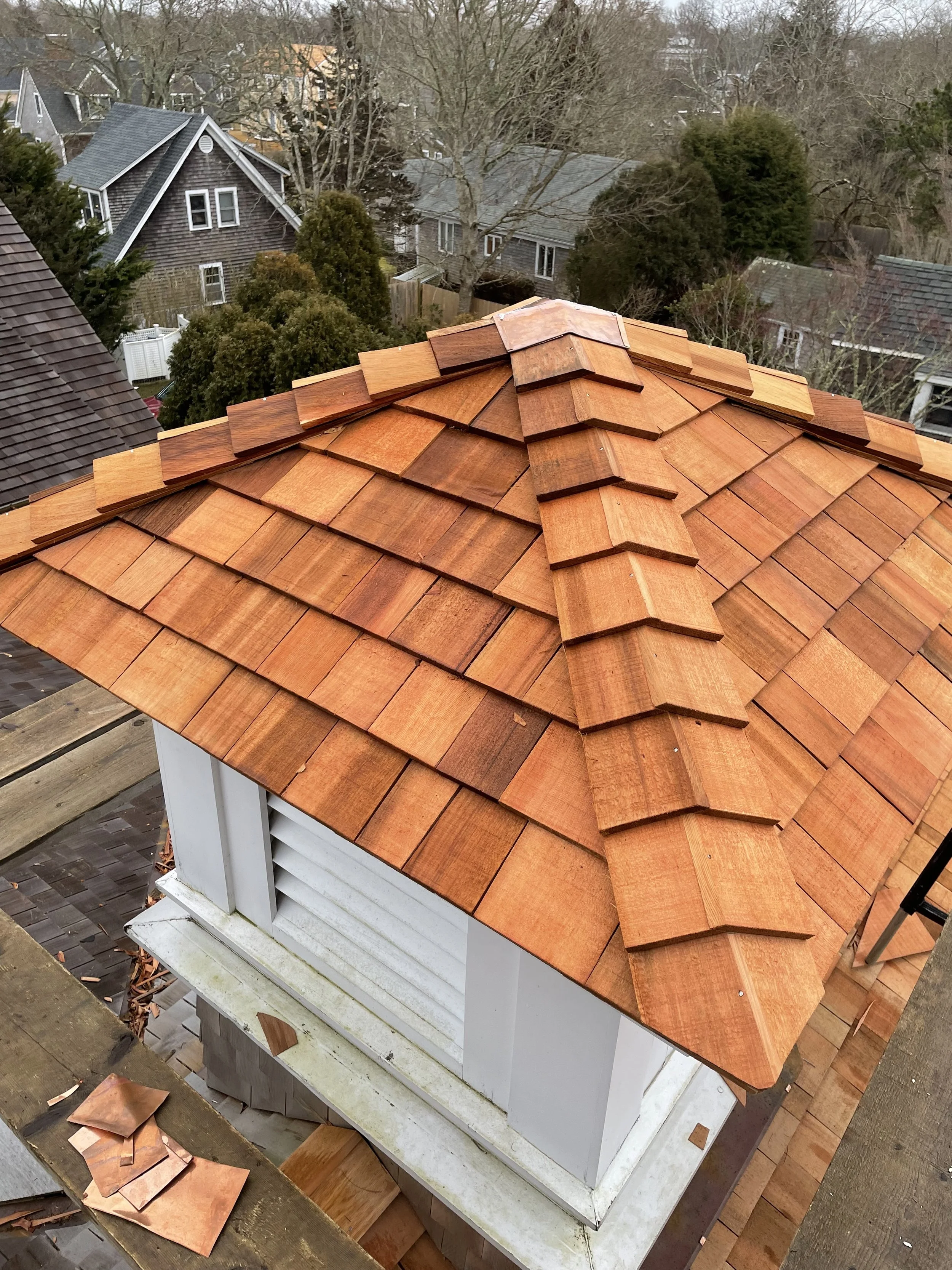 Close-up of a newly installed reddish-brown shingle roof on a white house, with a background of trees and neighboring houses.