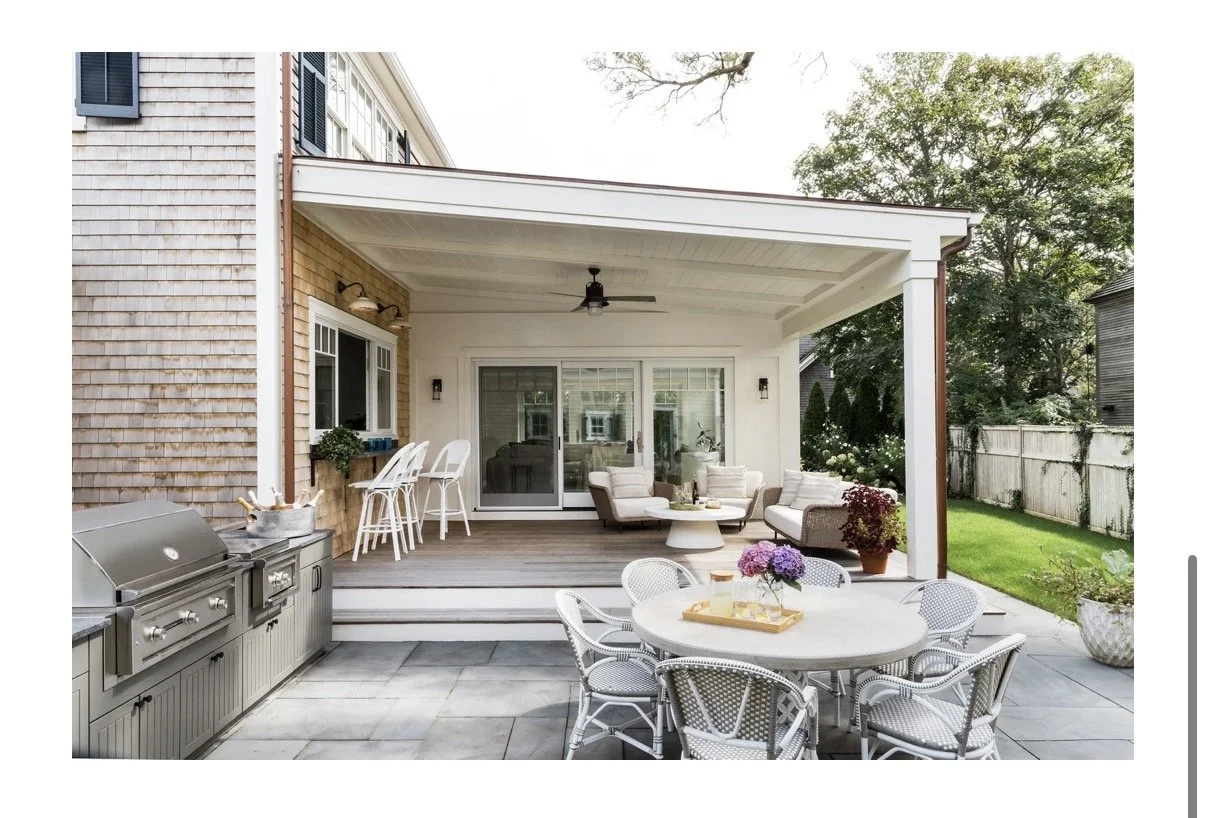 This image shows a backyard patio with outdoor furniture, including a round dining table with chairs and a sectional sofa with cushions under a covered porch. A grill is on the paved area, and a wooden fence surrounds the yard with grass and trees.
