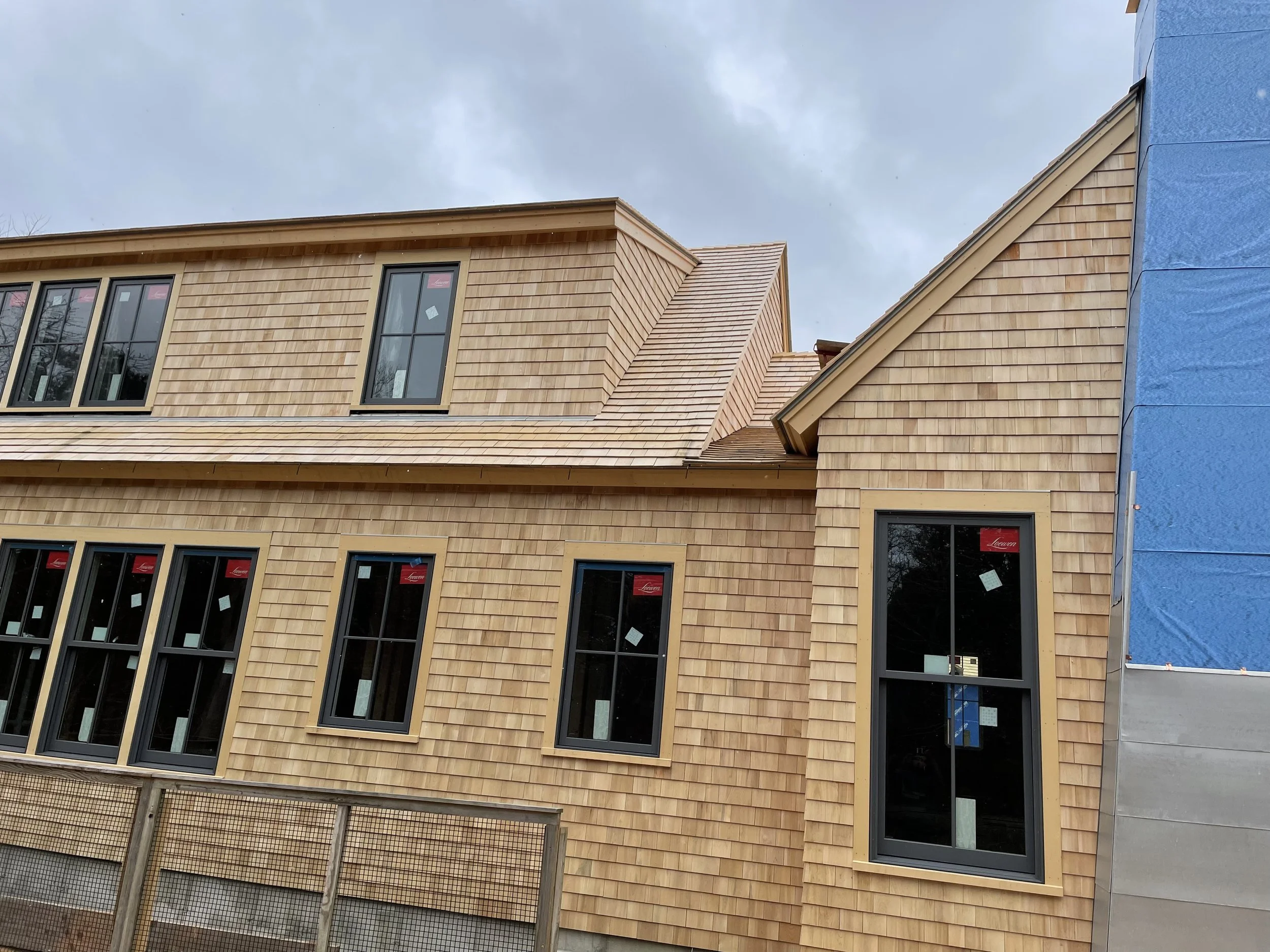 A building under construction with beige wooden siding, multiple windows, and a blue section of exterior sheathing visible on the right side against a cloudy sky.